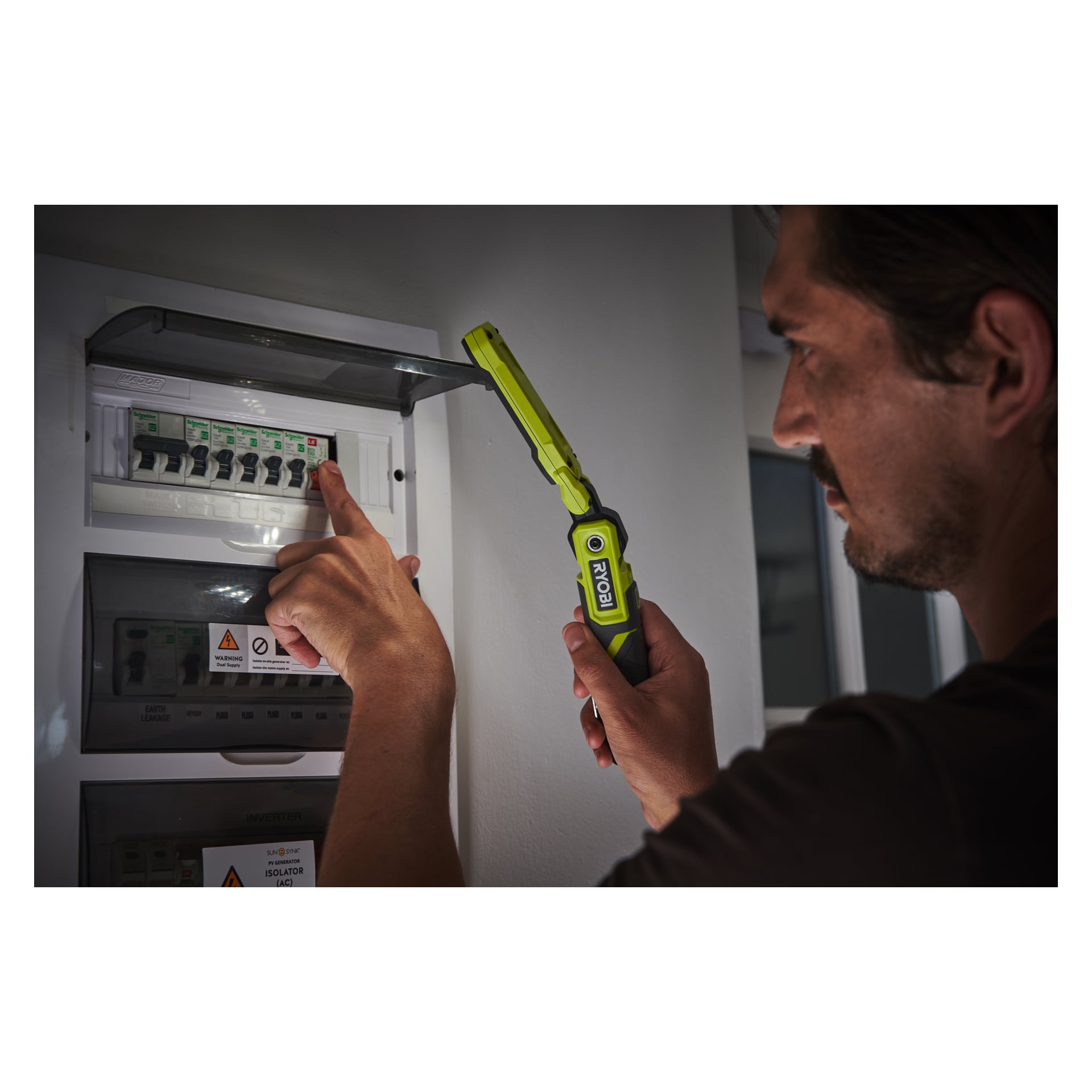 En man håller en grön Ryobi USB Lithium Work Lamp RLI4-0 nära en säkringspanel och riktar ljuset mot en av strömbrytarna. Säkringsboxen innehåller flera strömbrytare och varningsskyltar. Han är inomhus, och väggarna är vita.