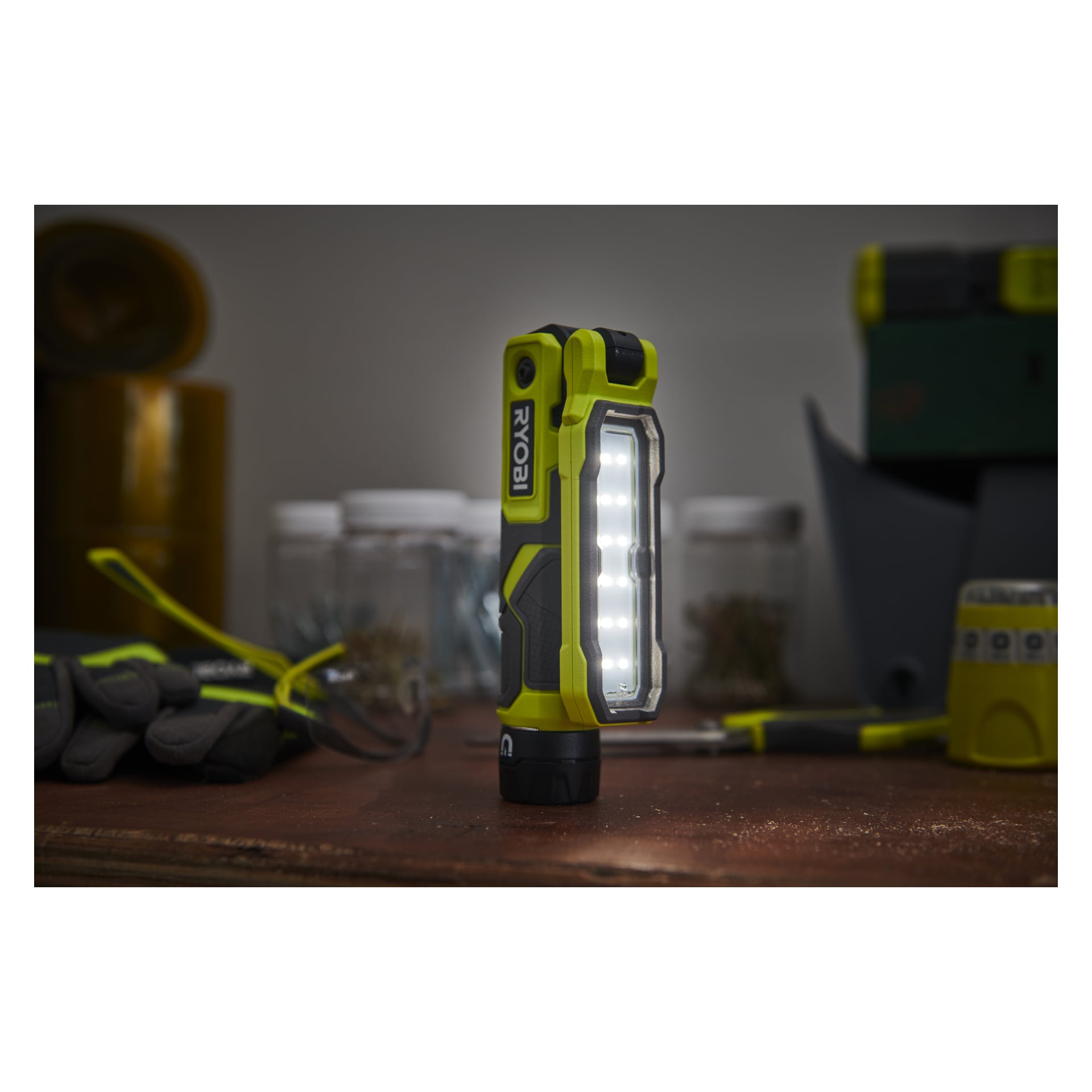En Ryobi USB Lithium Work Lamp RLI4-0 är placerad upprätt på en arbetsbänk och lyser upp arbetsytan. I närheten finns handskar, burkar och diverse verkstadsföremål.