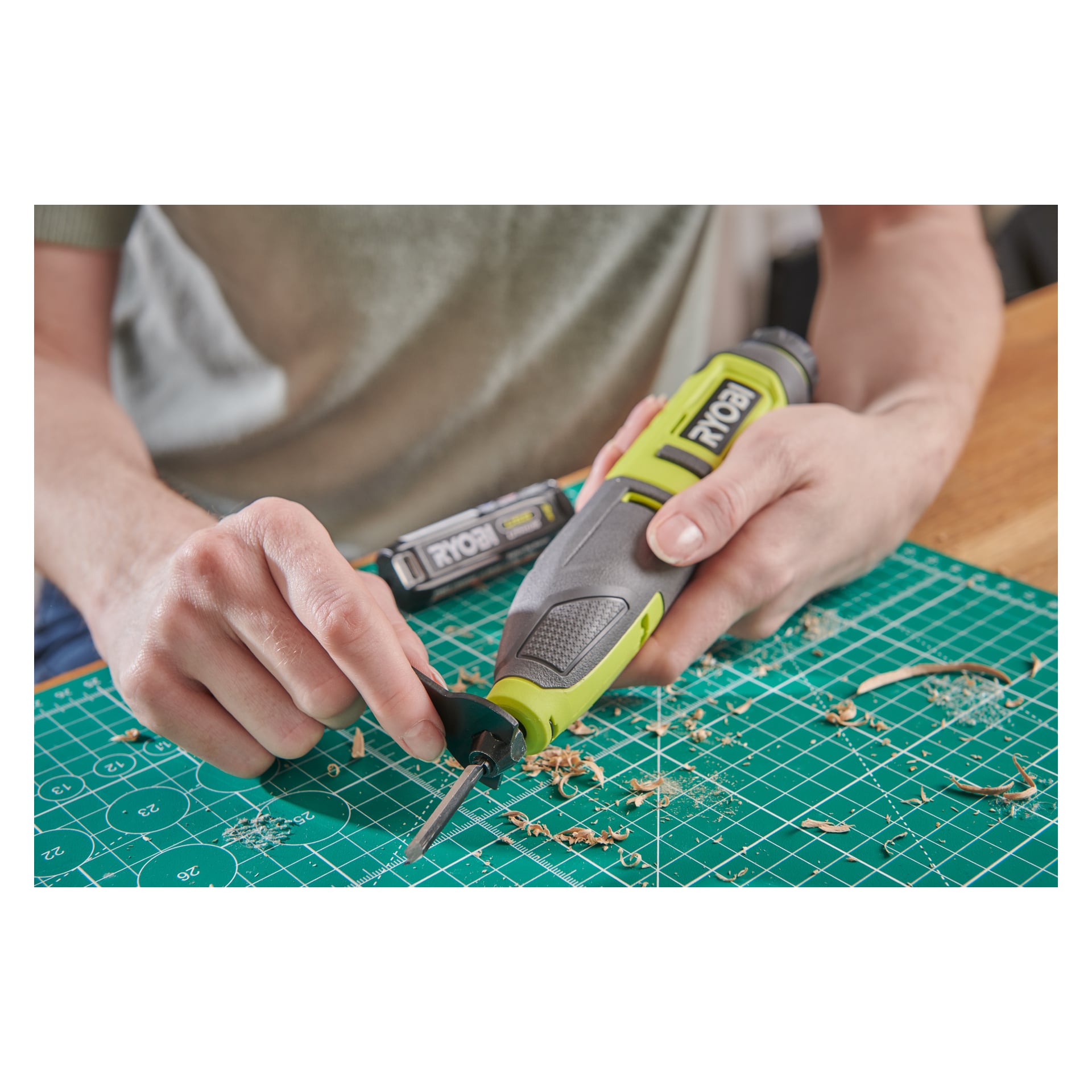 Człowiek starannie rzeźbi drewno zielono-czarnym dłutem Ryobi USB Lithium Power Power RPC4-0 na macie do cięcia. Pokazane są ręce prowadzące narzędzie do szczegółowej pracy, z wiórami drewnianymi i baterią Ryobi w pobliżu.