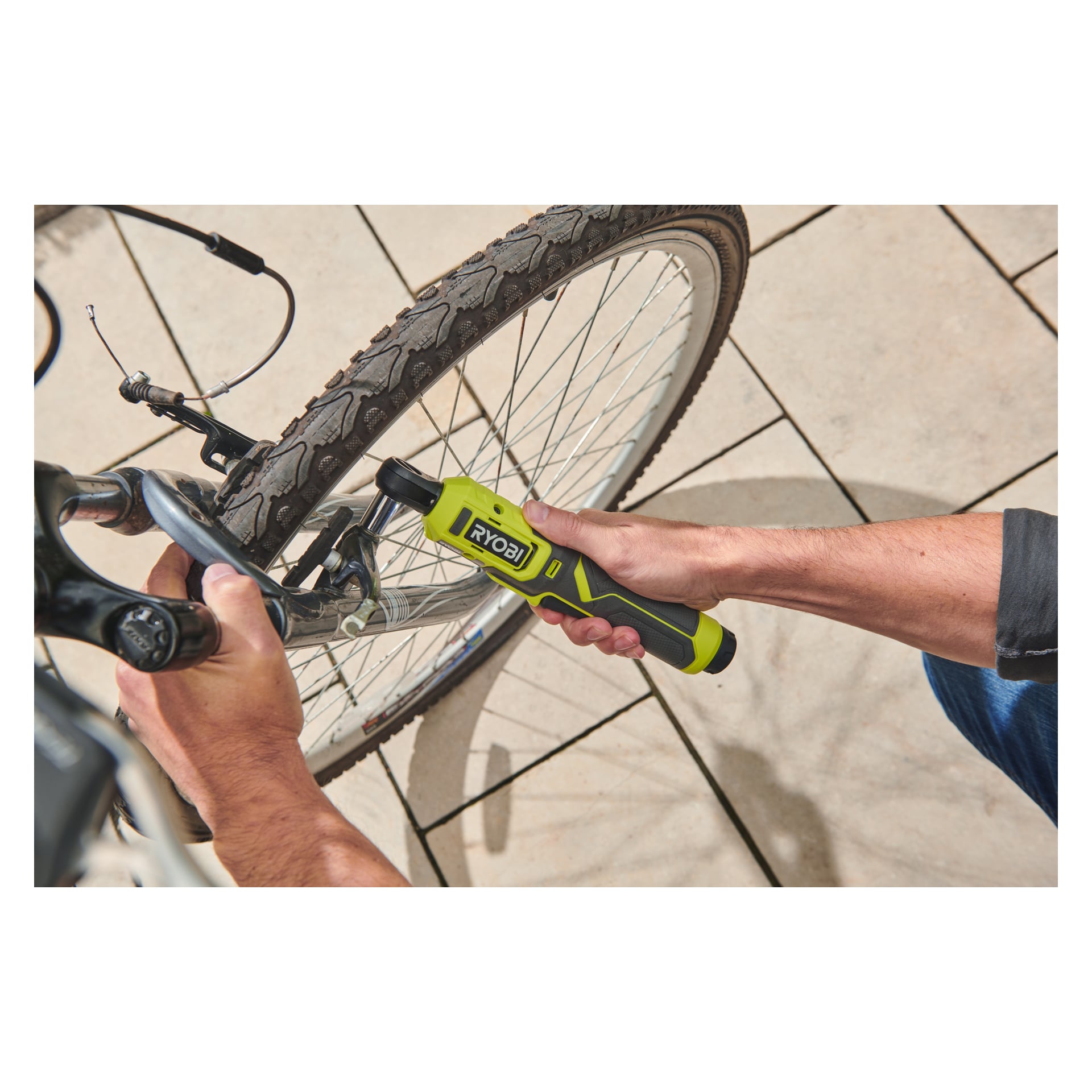 Någon använder en Ryobi USB litiumbatteridriven spärrnyckel RR14W4-0 för att fästa en bult på cykelns främre bromsmekanism. Cykeln står på en utomhus kaklad yta, och båda händerna greppar verktyget och bromsdelen.