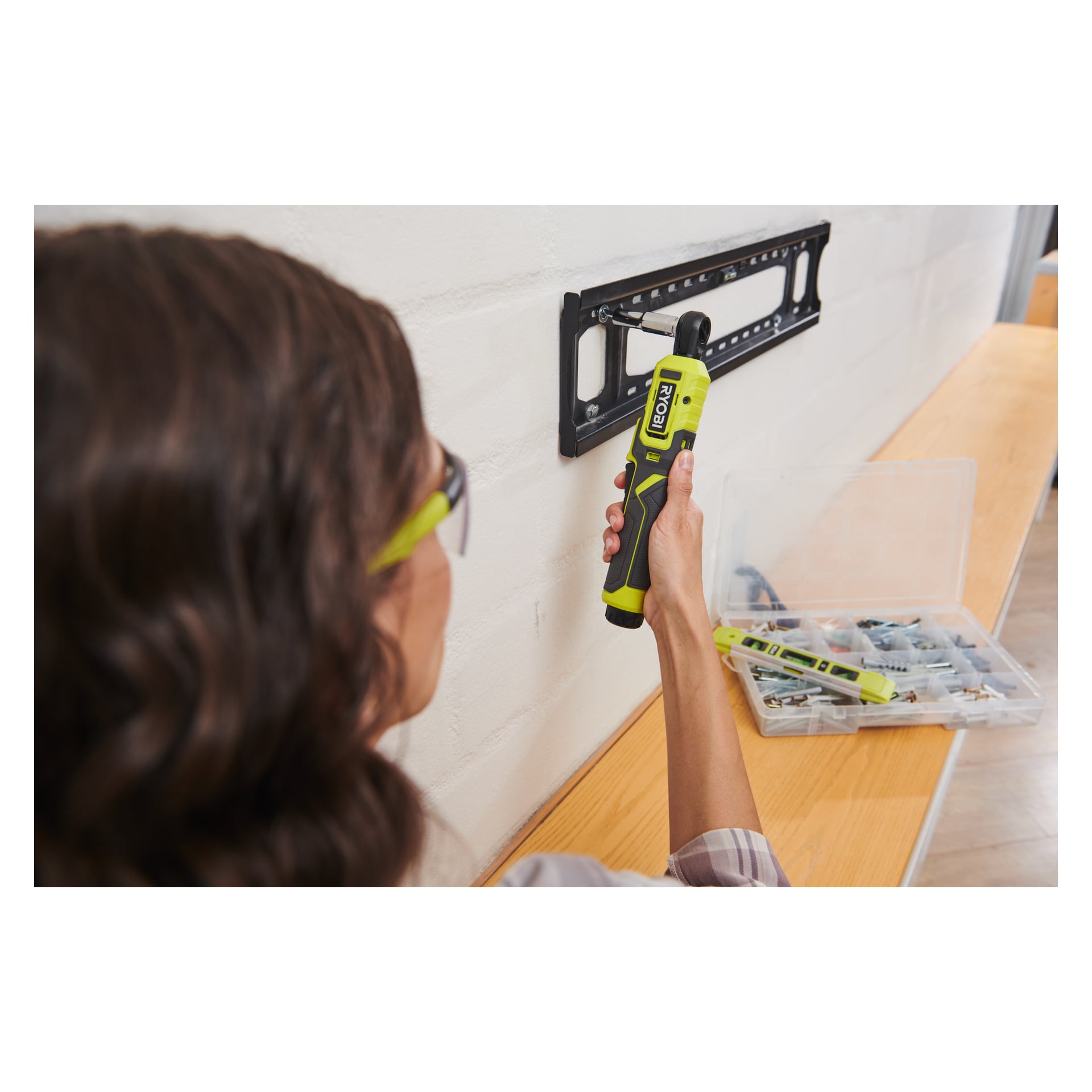 En person med skyddsglasögon använder en grön Ryobi USB litium-spärrnyckel RR14W4-0 för att fästa ett TV-väggfäste på en vit vägg. På ett träbord i närheten står en genomskinlig plastlåda fylld med skruvar och verktyg.