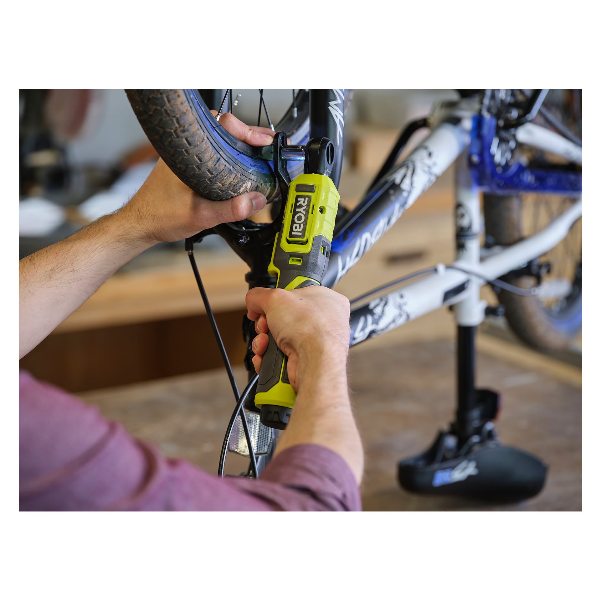 Någon använder en Ryobi USB Lithium Battery spärrnyckel RR14W4-0 medan man arbetar på framhjulet på en upp-och-nedvänd cykel som står på en arbetsbänk i en verkstadmiljö.