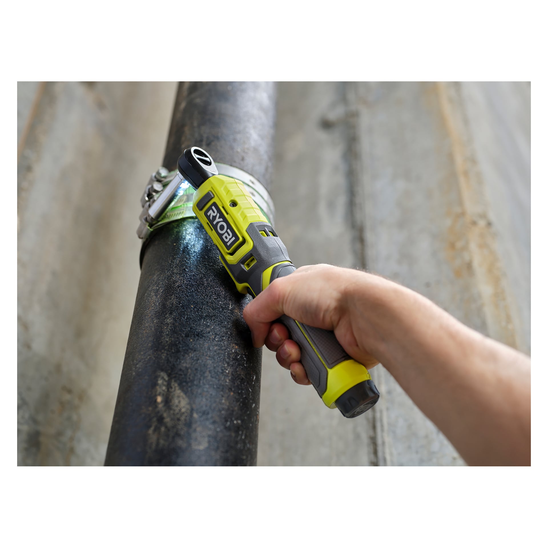 Någon drar åt en klämma runt ett stort svart rör med hjälp av en Ryobi USB litiumbatteridriven spärrnyckel RR14W4-0 i en industri- eller byggmiljö med betongytor.