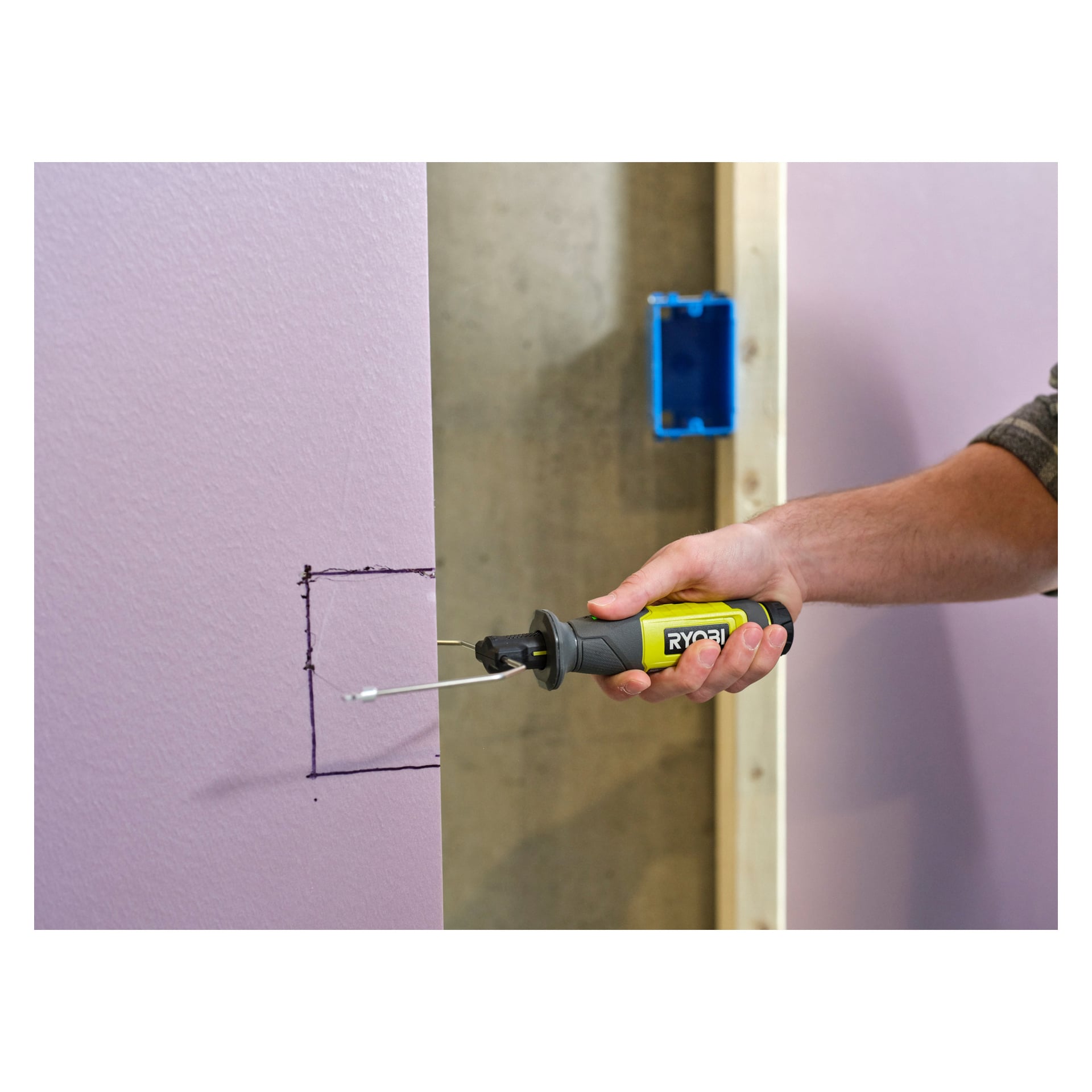 Du bruker en Ryobi USB Lithium Battery hot cutter RHC4-0 for å skjære ut en markert firkant fra et lilla gipspanel. I bakgrunnen er det en trestender og en elektrisk boks.