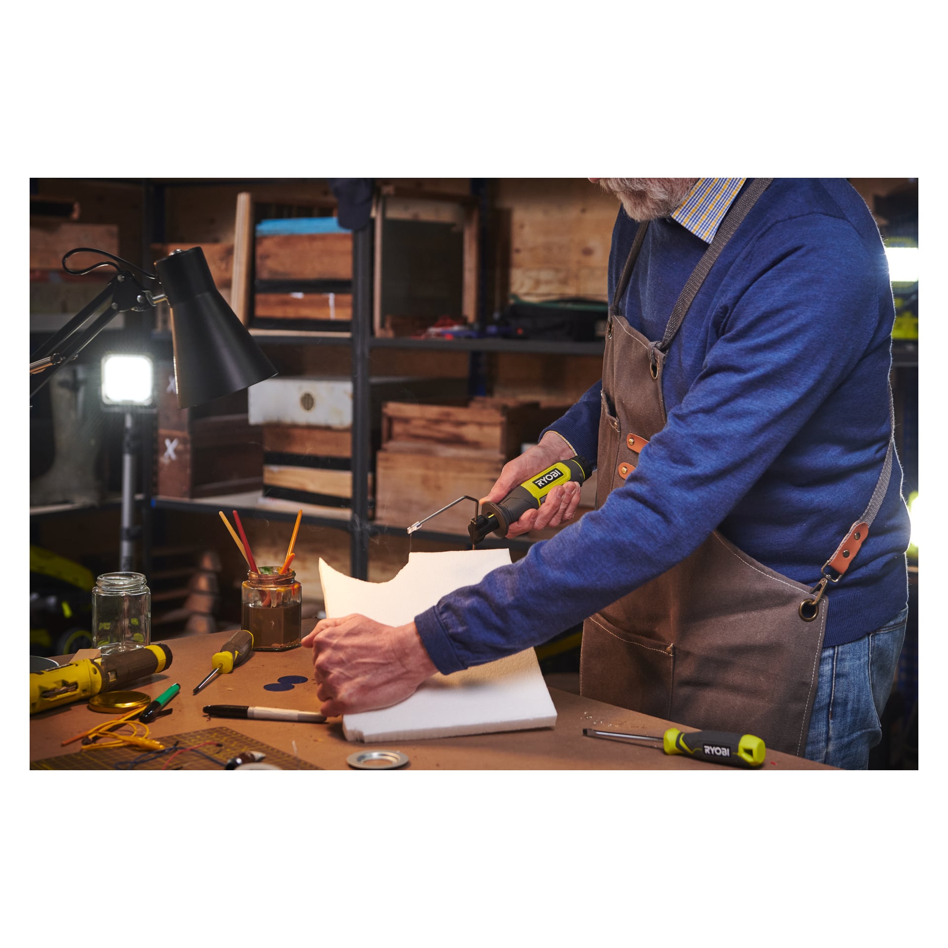 En person i blå genser og forkle bruker Ryobi USB Lithium Battery-drevne hot cutter RHC4-0 til å skjære gjennom et hvitt skumark på en arbeidsbenk, omgitt av verktøy, pensler og diverse verkstedmateriell på bordet og hyllene.