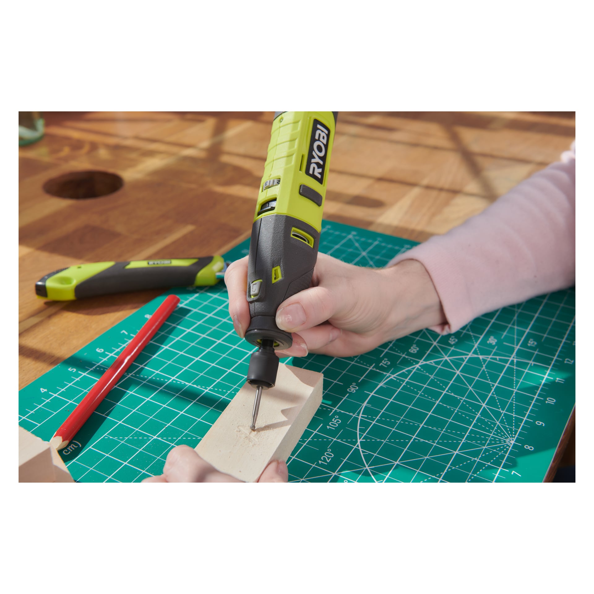 Någon använder ett Ryobi USB litiumbatteridrivet RRT4-120GA15 multiverktyg, där man borrar i en liten träbit som placeras på en grön skärmatta. I närheten på träbordet ligger en röd penna och en grön hobbykniv.