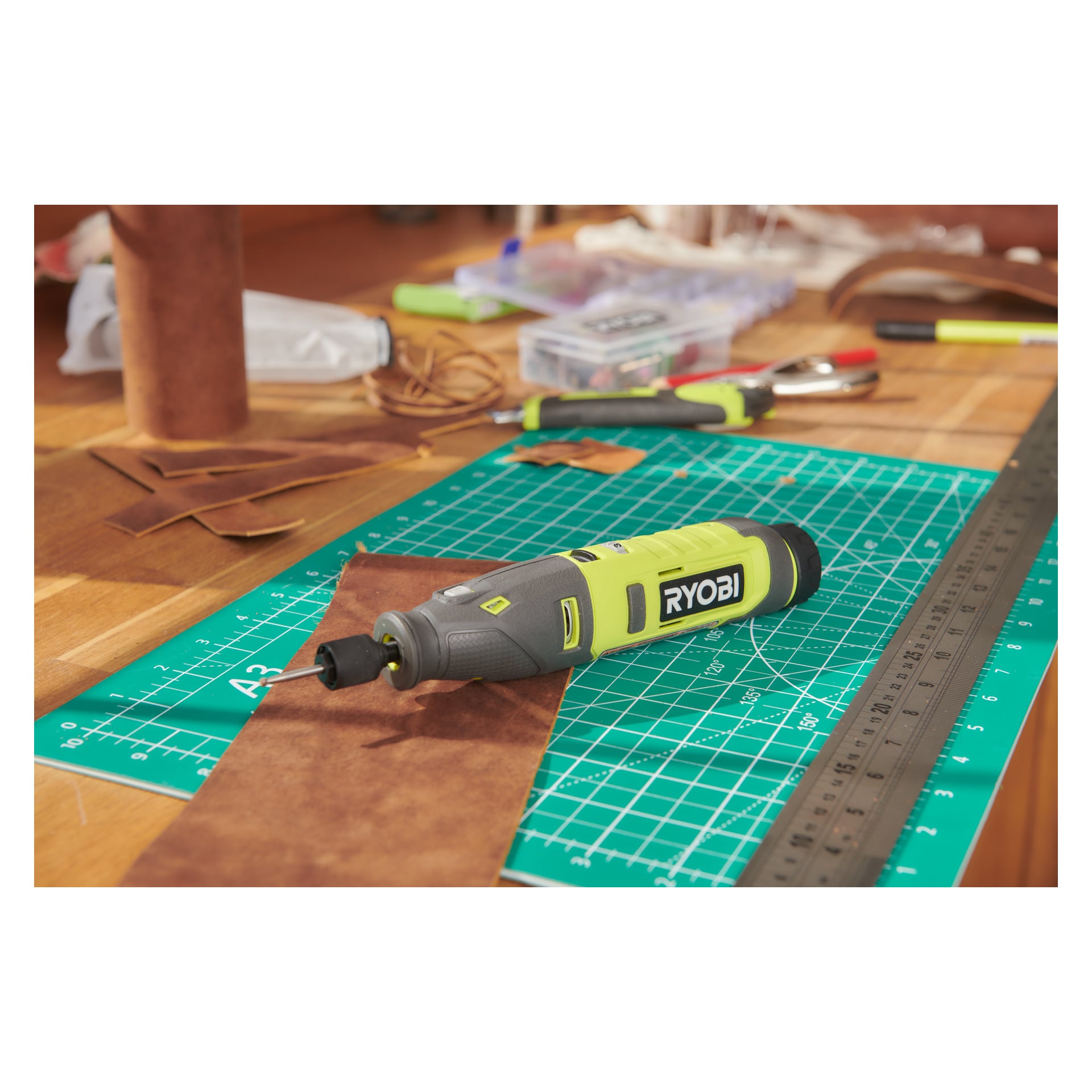 En Ryobi USB Lithium Battery-driven multiverktygs-RRT4-120GA15 (19 delar) ligger på en grön skärmatta bredvid en metalllinjal och bruna läderdelar, medan diverse verktyg och förnödenheter är utspridda över den träyta i bakgrunden.