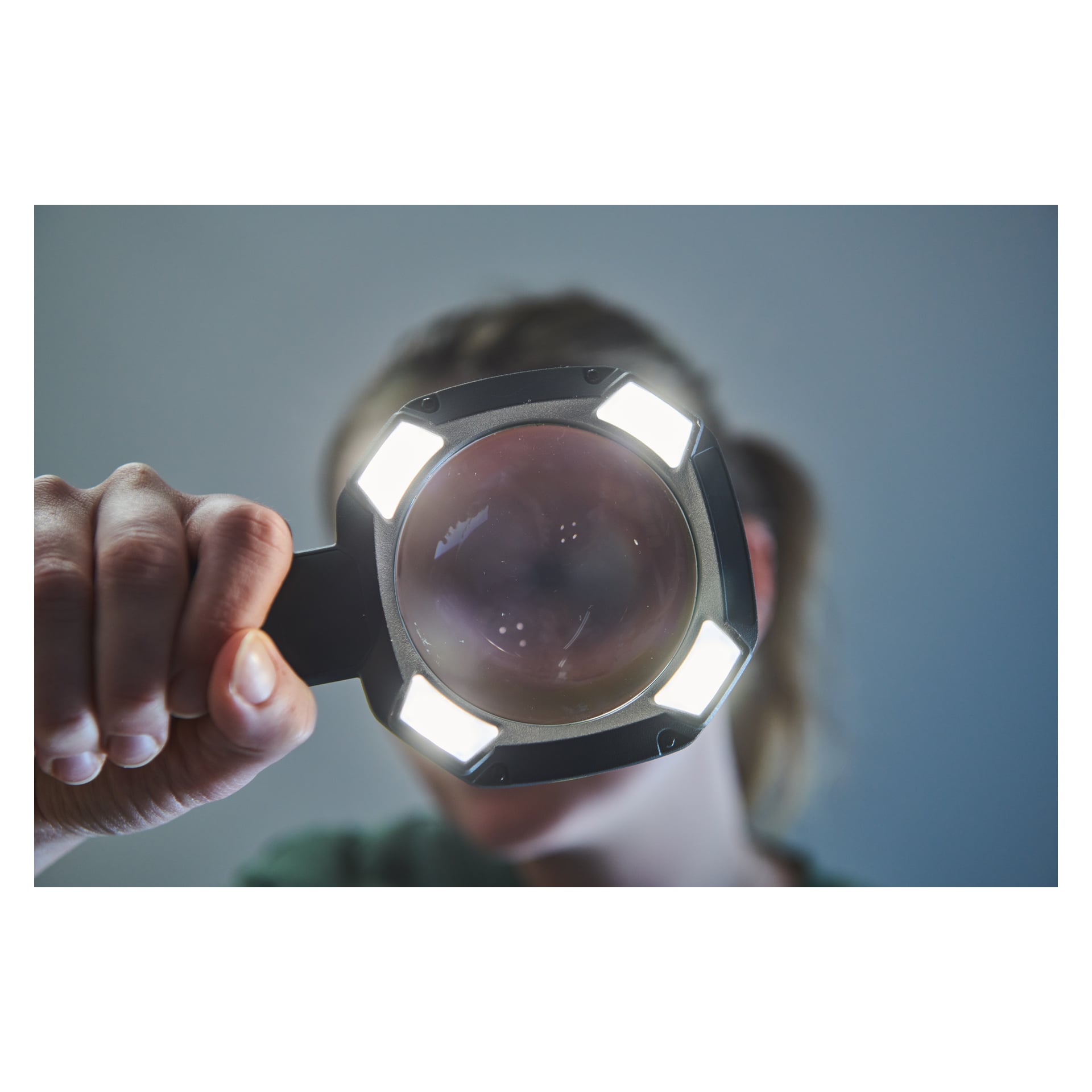 En person håller ett Ryobi USB litiumförstoringsglas med lampa RML4-0 nära kameran, med sina fyra LED-lampor som lyser genom den stora linsen och delvis täcker ansiktet. Deras hand syns greppa handtaget på vänster sida av scenen.