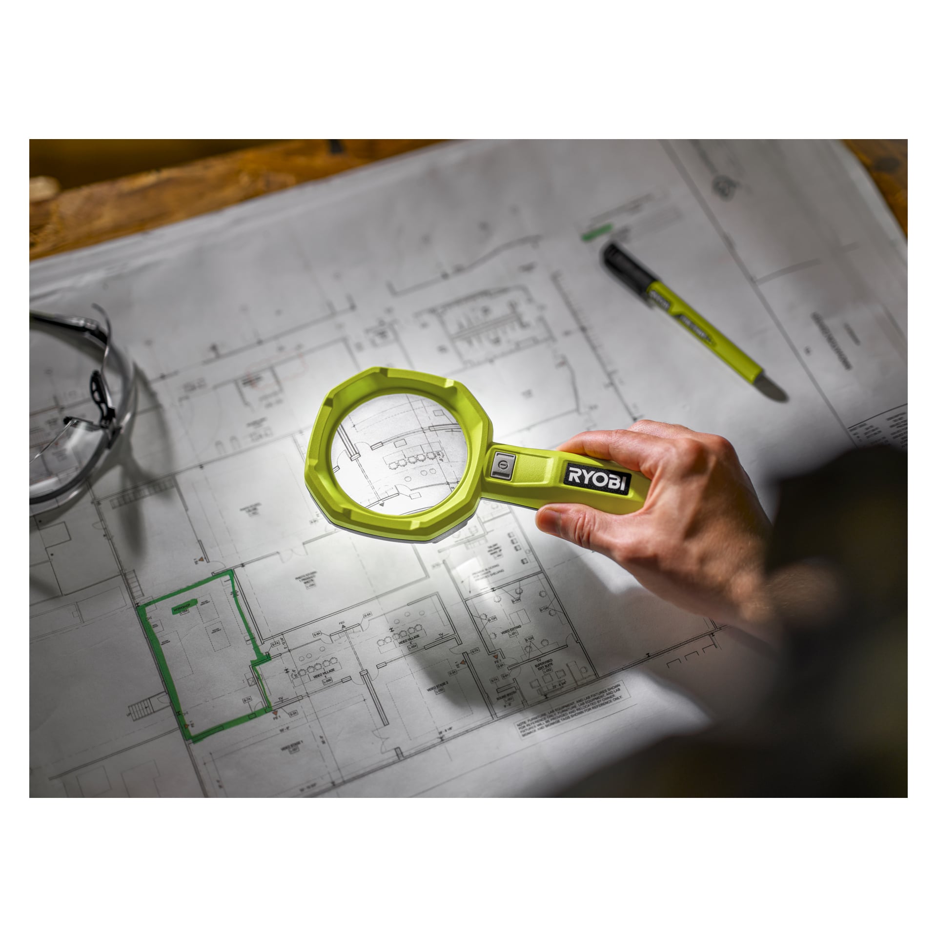 En hand håller ett grönt Ryobi USB Lithium förstoringsglas med lampa RML4-0 för att inspektera en teknisk ritning utlagd på ett bord, medan en del av en penna och ett par läsglasögon vilar i närheten på ritningen.