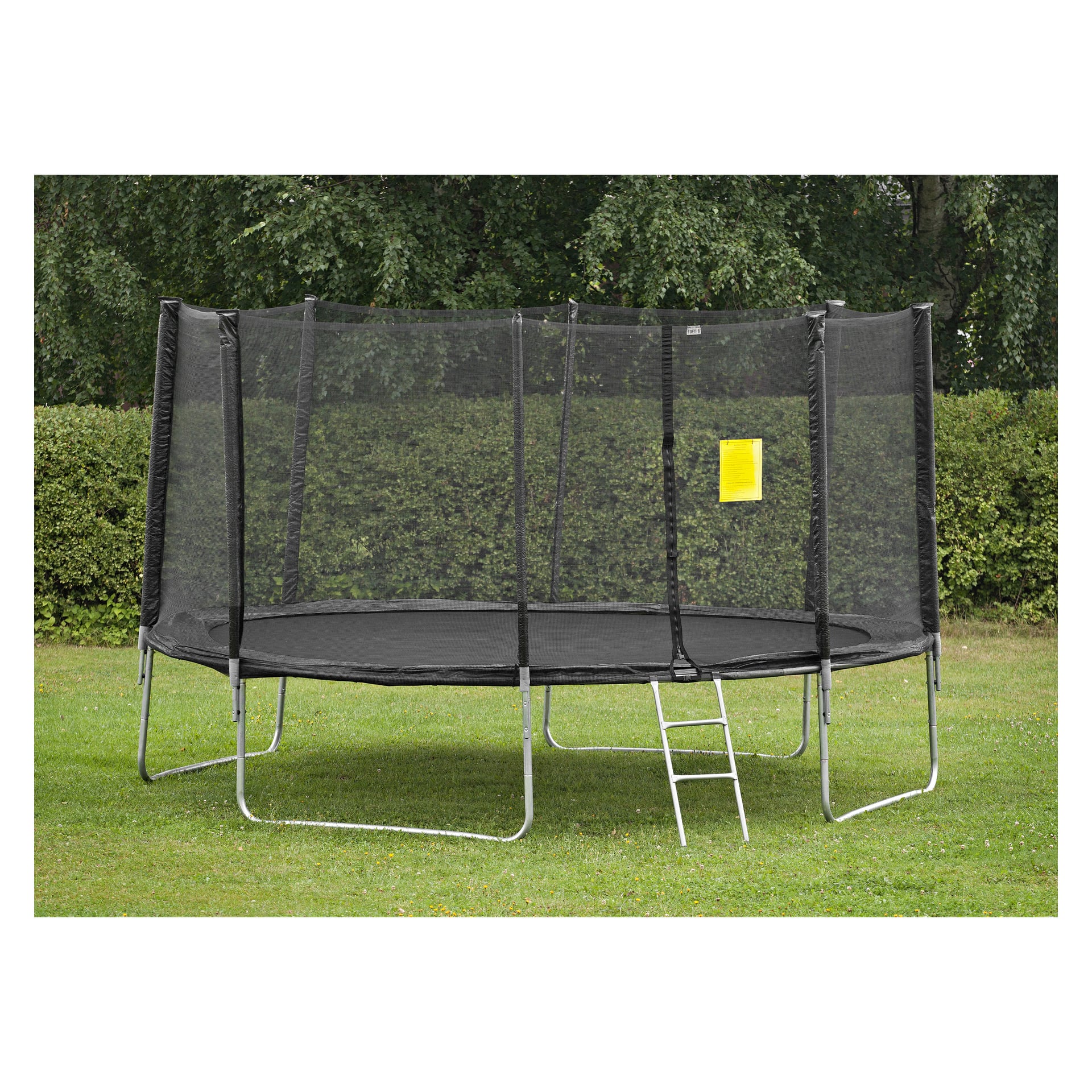 En rund svart Kayoba-trampolin med säkerhetsnät och metallstege är uppställd på gräsbevuxen mark i en trädgård. En gul skylt hänger från nätet, med buskar och träd synliga bakom.