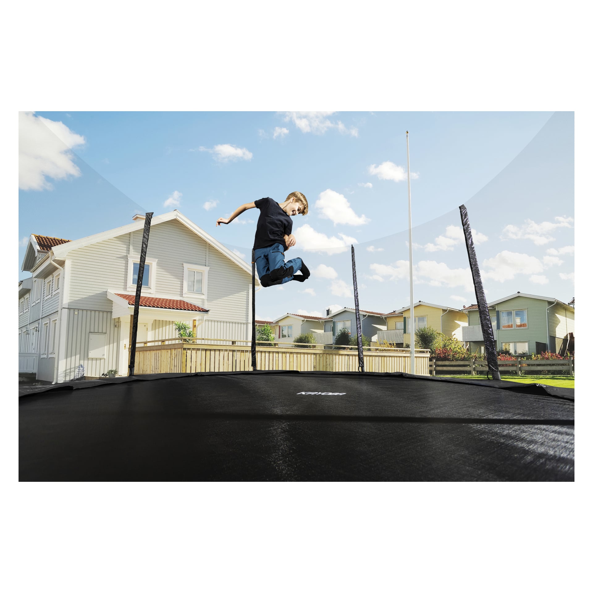 En pojke klädd i svart skjorta och blå byxor hoppar på en utomhus Kayoba-trampolin instängd av skyddsnät; Bakom honom finns vita hus och en himmel fylld av moln.
