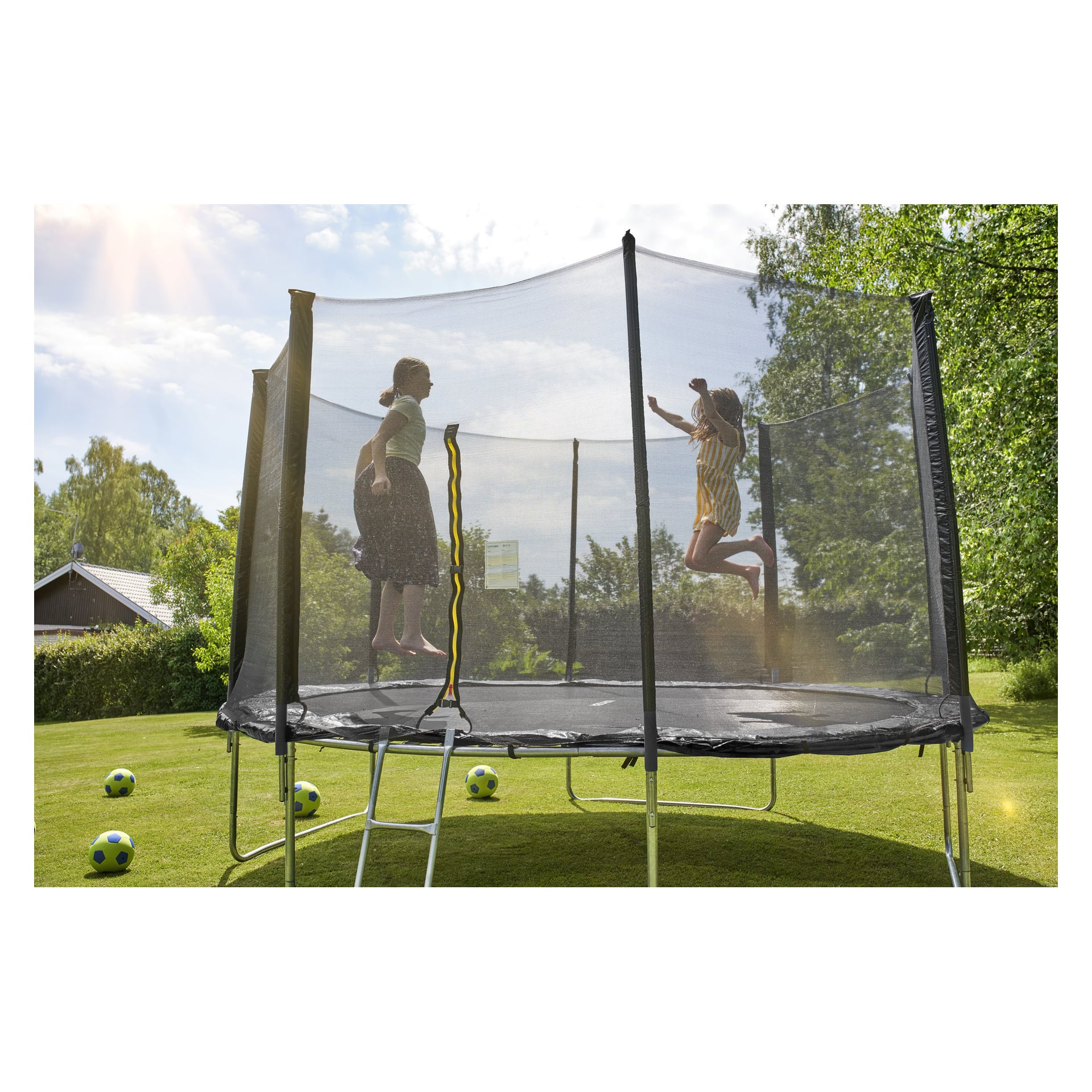 Två barn leker på en Kayoba-trampolin utrustad med skyddsnät i en solig, gräsbevuxen trädgård. Flera fotbollar vilar på gräset i närheten. I bakgrunden finns träd och ett hus.