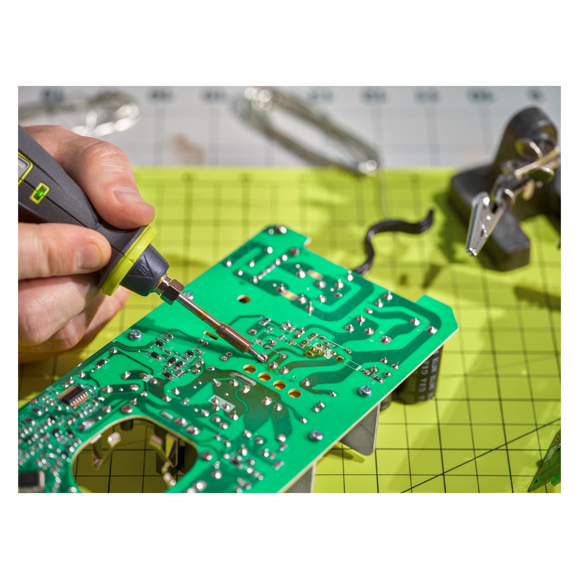 Ktoś pracuje nad zielonym obwodem płyty elektronicznej, używając lutownicy Ryobi USB zasilanej baterią litową RSI4-0. Różne metalowe narzędzia i komponenty elektroniczne są rozrzucone na powierzchni o żółtym wzorze siatki.