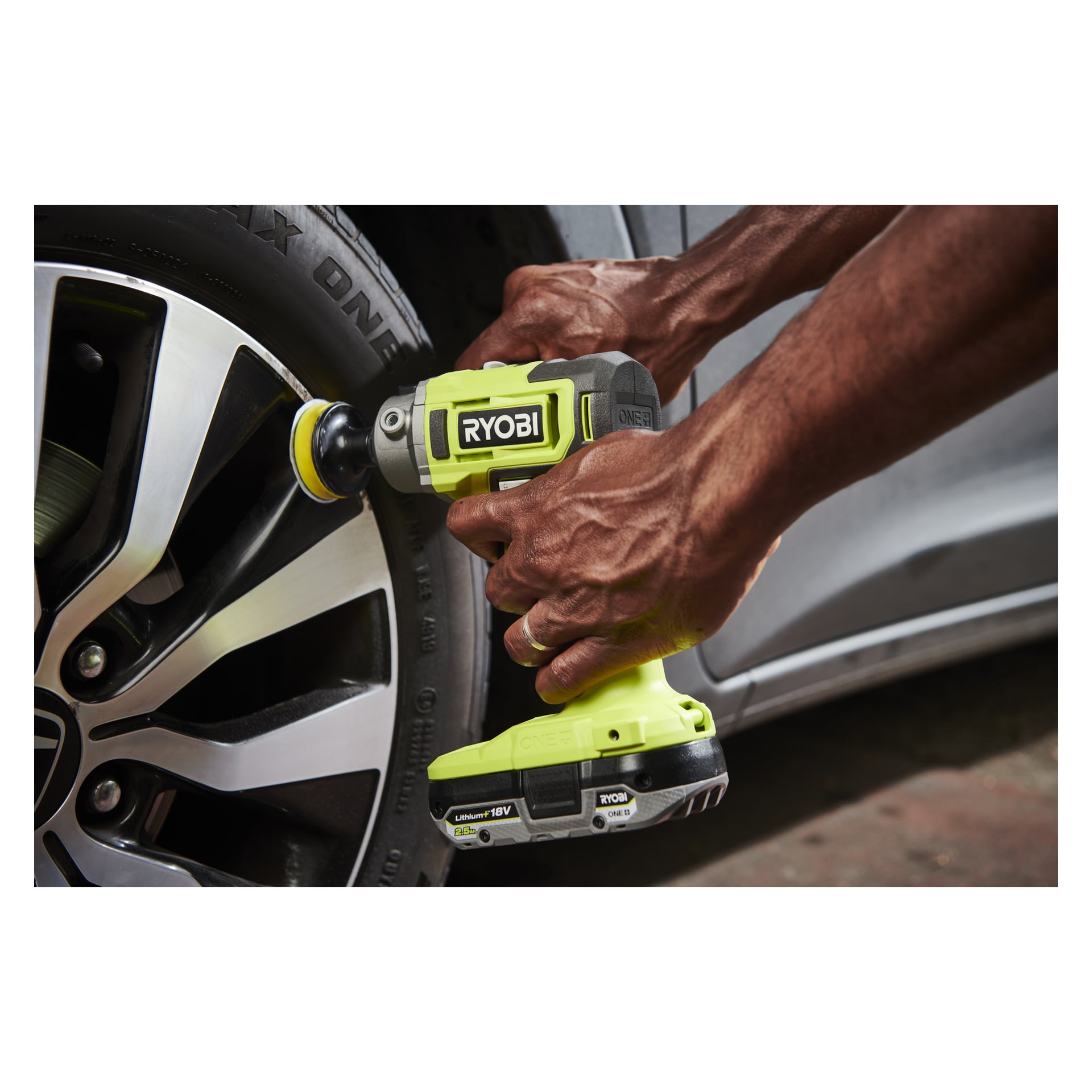 Någon håller i en Ryobi One+ Polisher/detaljslipare RDP18-0 utrustad med en borste för att rengöra kanten på ett bildäck, med fordonets hjul och däck synliga i bakgrunden.