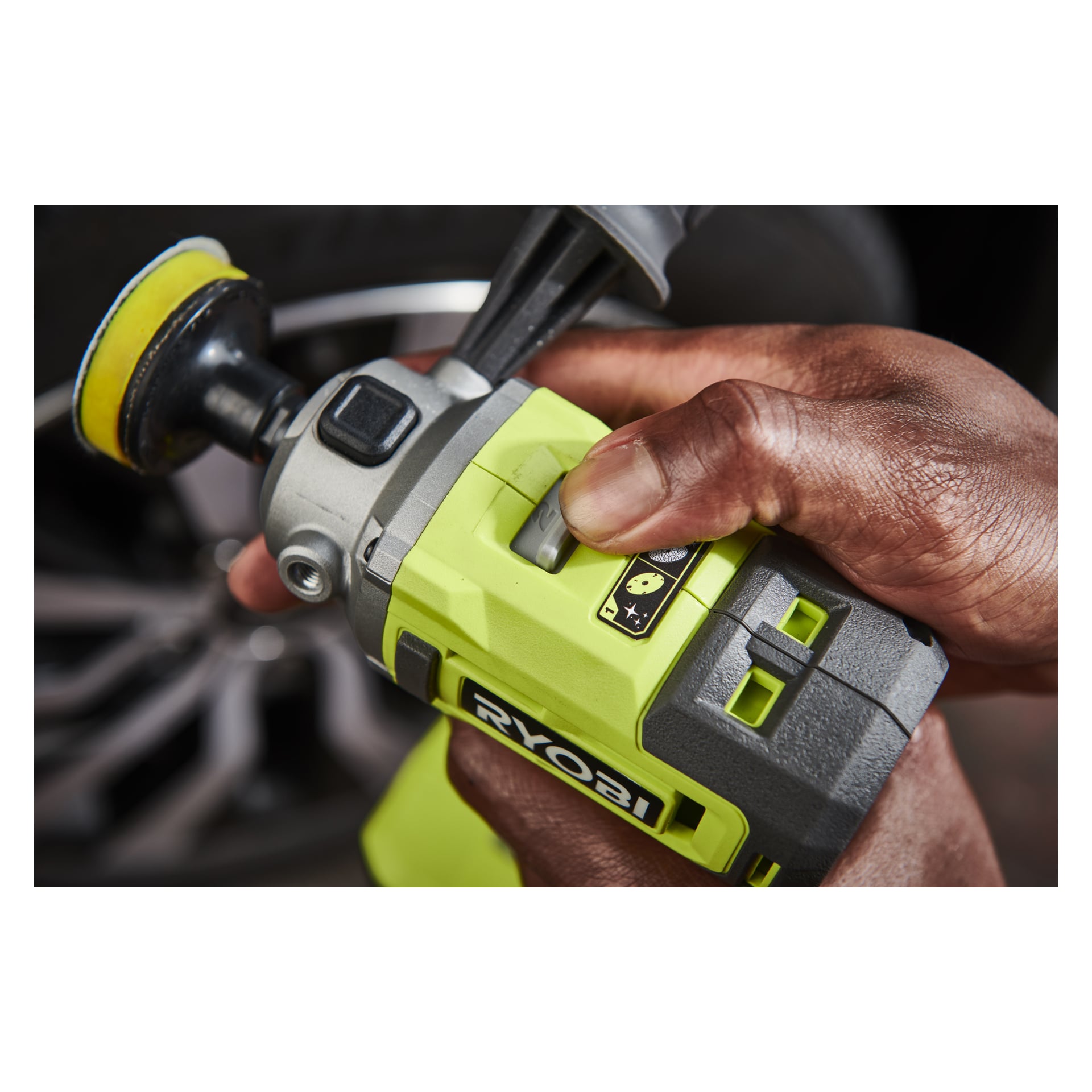 En hand vrider snabbratten på en grön Ryobi One+ Polisher/detaljslipare RDP18-0, och visar dess svarta och gula fäste nära ett suddigt bilhjul i bakgrunden.