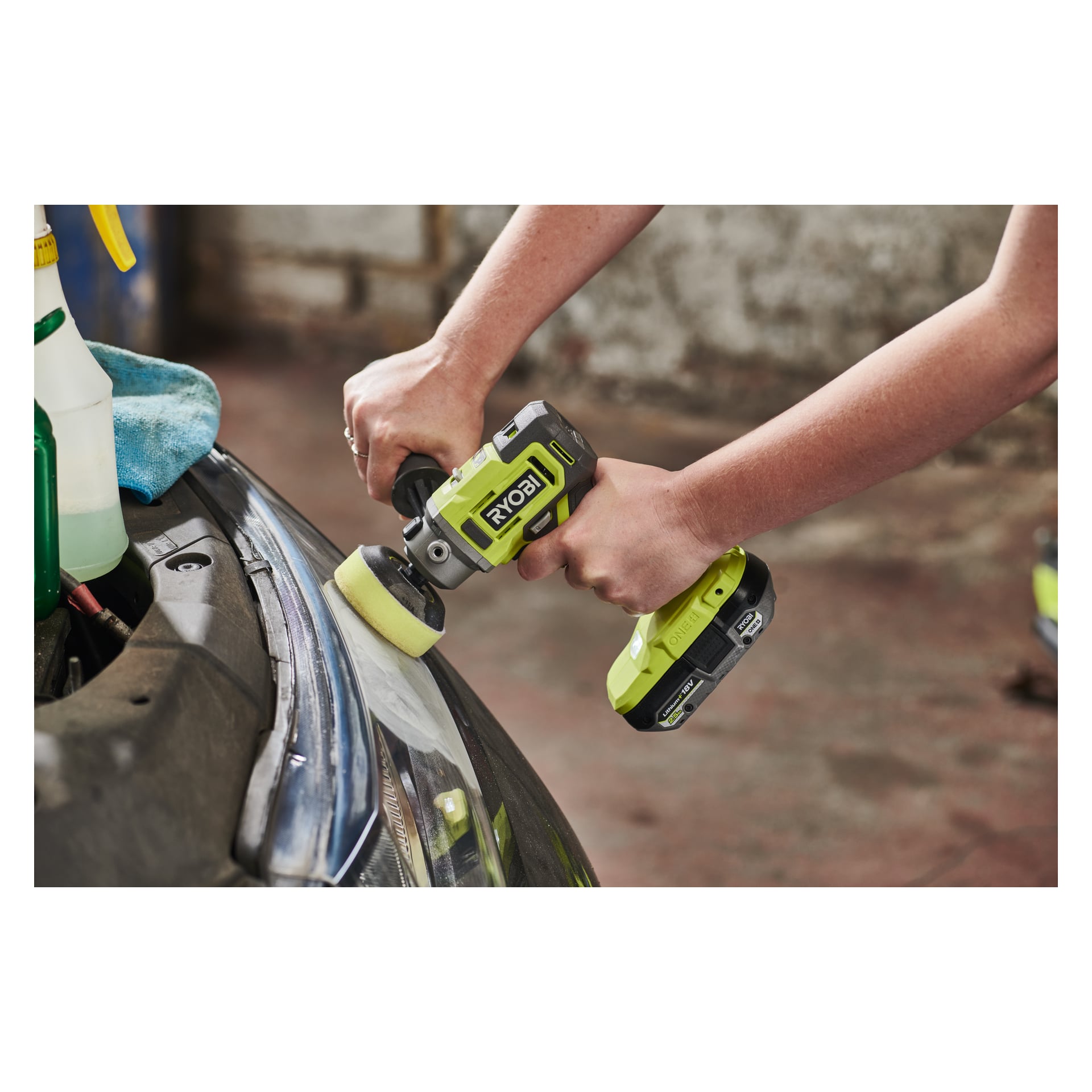 En person polerar en bils strålkastare med en Ryobi One+ Polisher/detaljslipare RDP18-0 utrustad med en gul pad. En sprayflaska och en blå handduk står på bilens kant, medan scenen utspelar sig i ett garage.