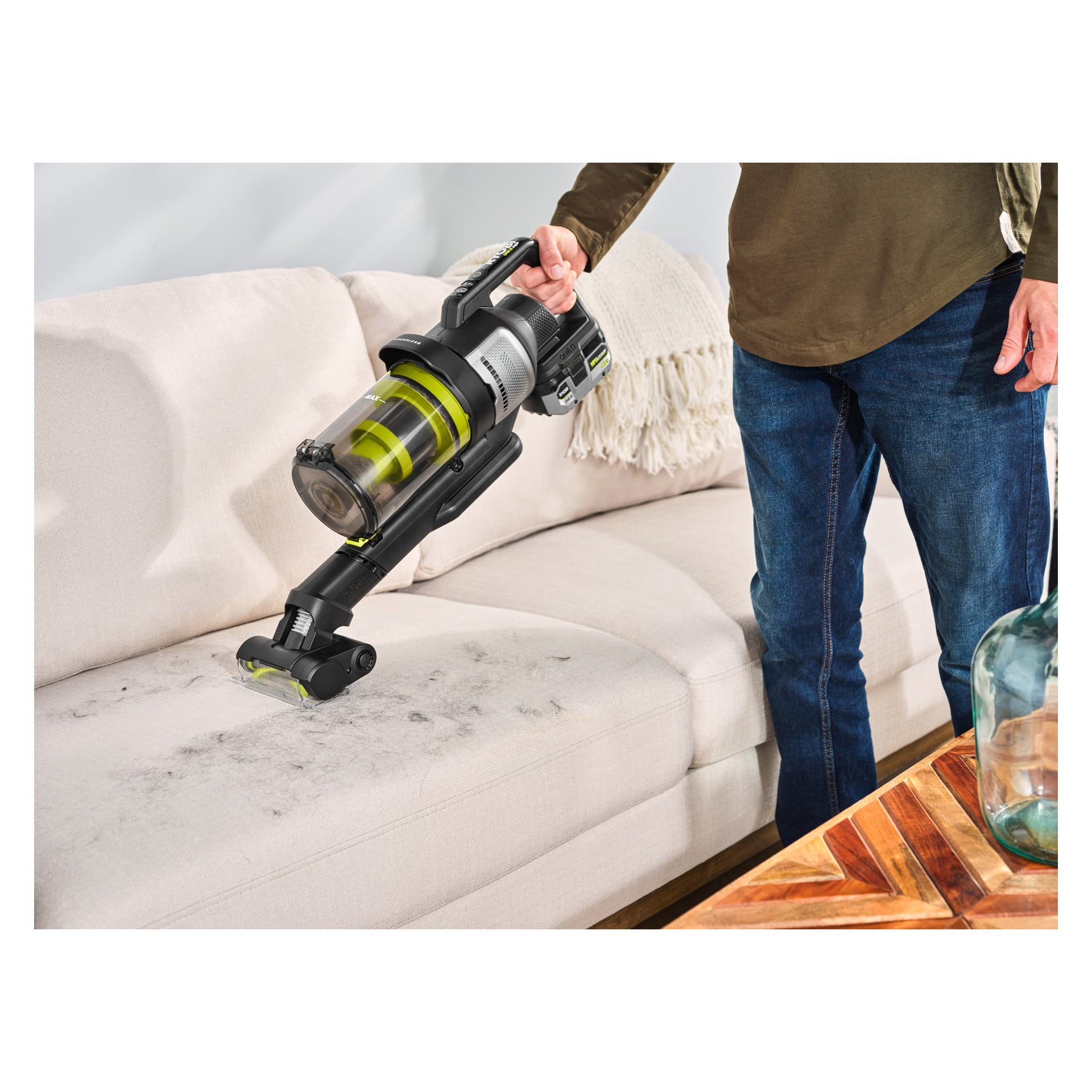 Någon rengör djurhår och smuts från en ljusfärgad soffa med Ryobi One+ Upright dammsugaren premium RASV18BL-1X40G, som riktar in sig på synligt skräp. I förgrunden kan man också se en del av ett träbord och lite glasdekoration.