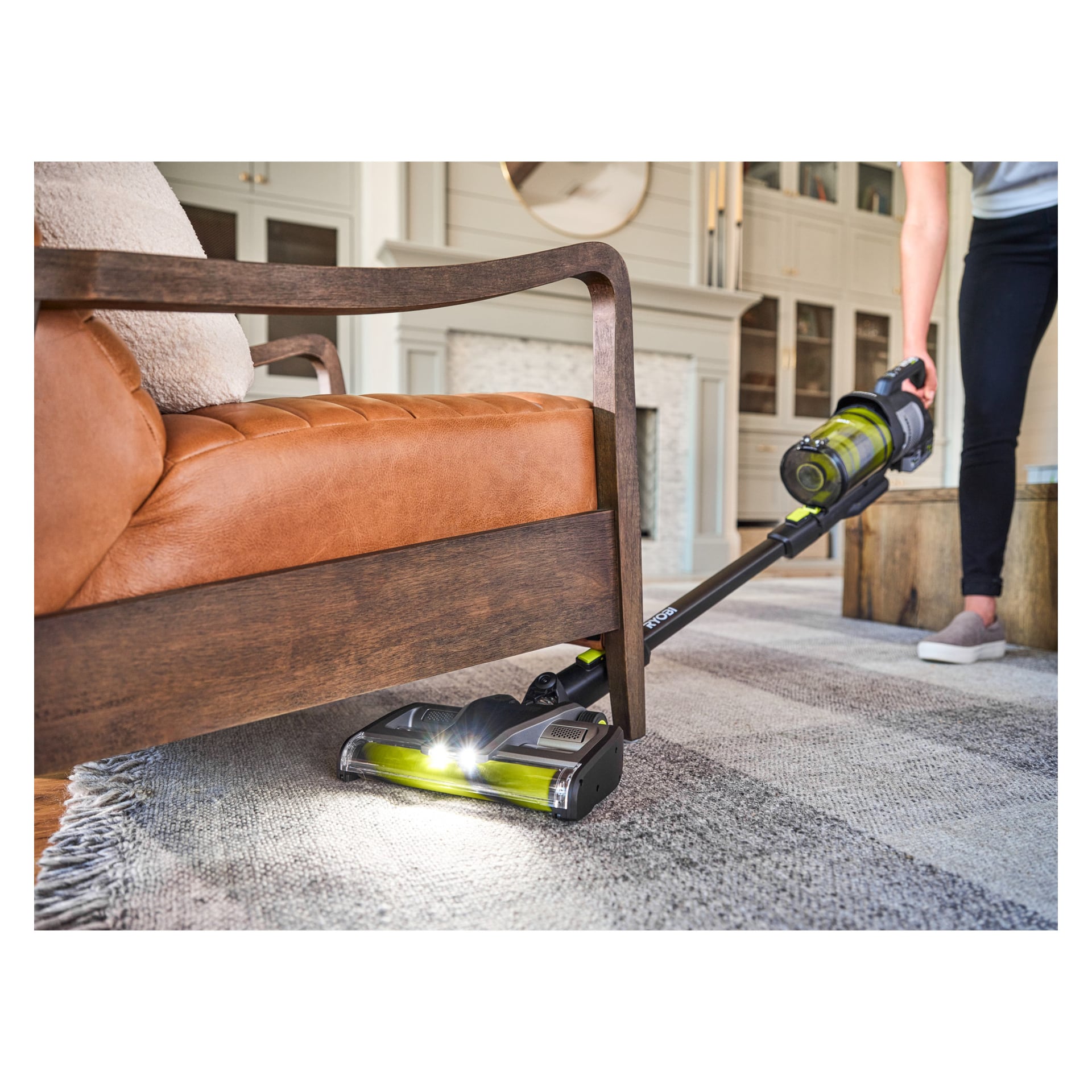 Man använder Ryobi One+ Upright Vacuum Cleaner Premium RASV18BL-1X40G för att rengöra under en brun soffa i ett vardagsrum med matta, trämöbler och inbyggda hyllor.
