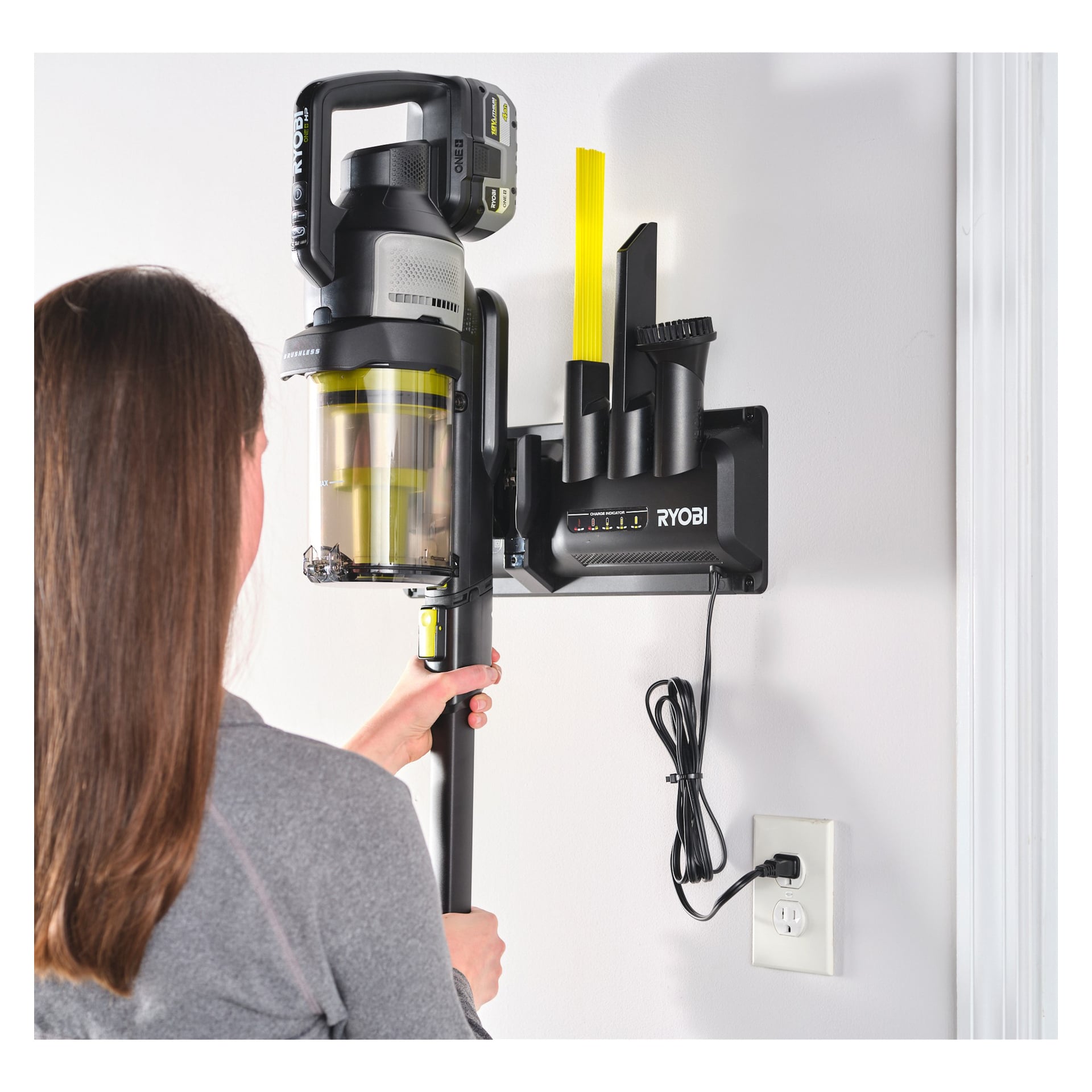 Man kopplar en Ryobi One+ Upright dammsugarmodell RASV18BL-1X40G till en väggladdningsstation med tillbehörshållare och kopplar strömkabeln till ett uttag.