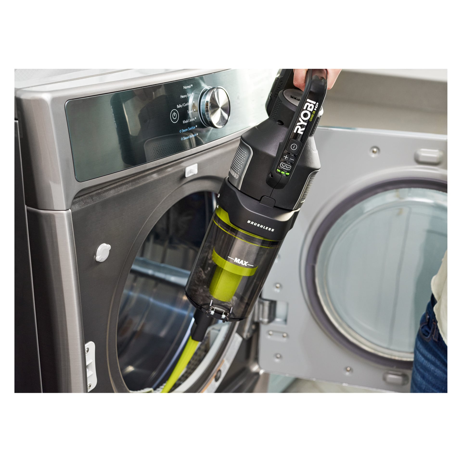 Någon rengör insidan av en frontmatad torktumlare med en Ryobi One+ Upright dammsugare premium RASV18BL-1X40G. Torktumlarens dörr är öppen och dammbehållaren på dammsugaren syns. Miljön verkar vara ett tvättrum.
