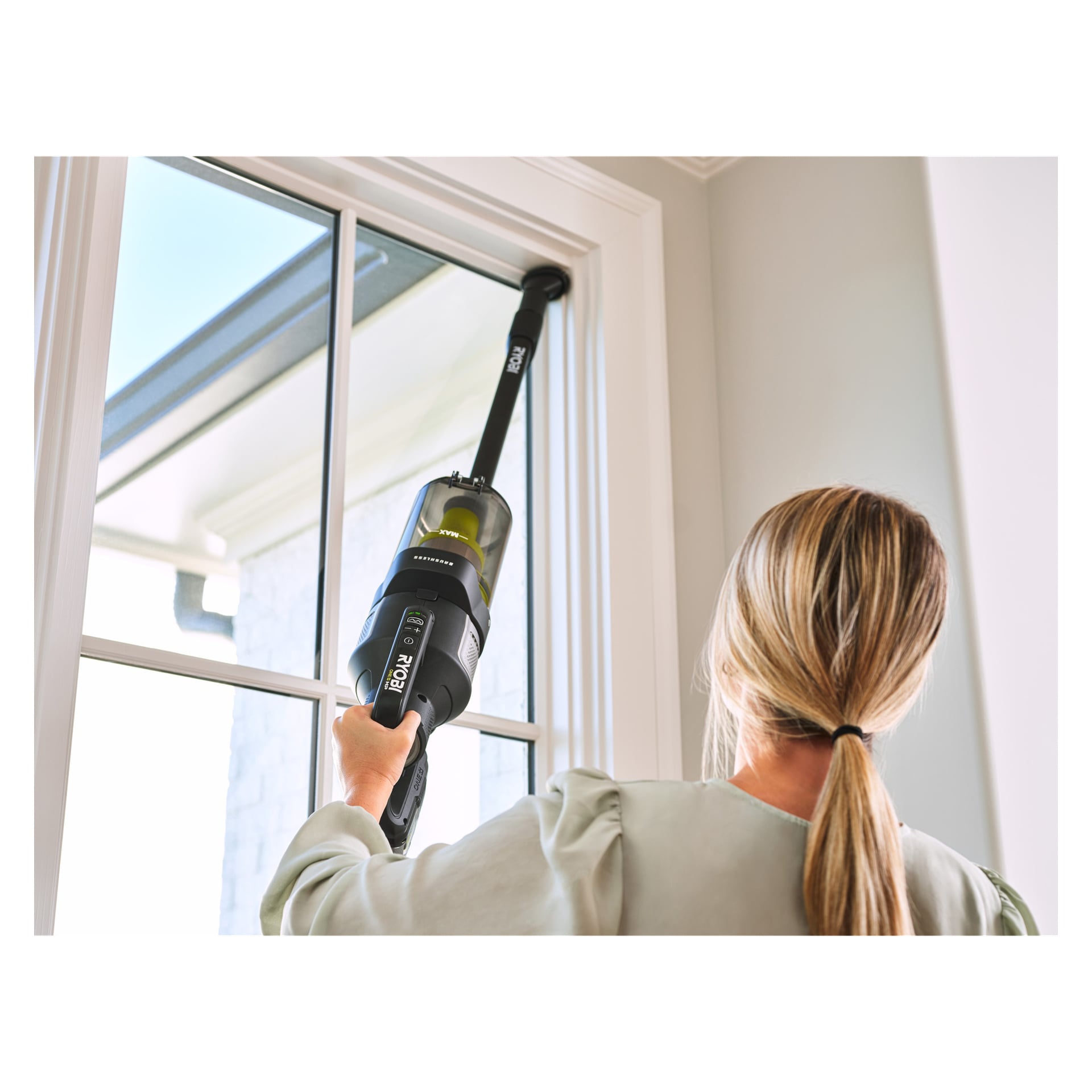Man rengör den övre kanten av ett fönster inne i ett väl upplyst rum genom att hålla upp Ryobi One+ Upright dammsugaren premium RASV18BL-1X40G med förlängningsanordning för att nå in i hörnet.