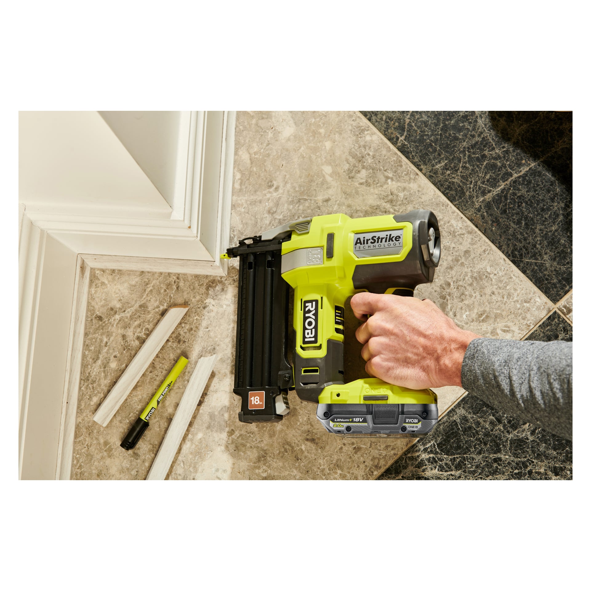 Joku käyttää Ryobi One+ -paristonaulapistoolia (R18GN18-1C20G), kiinnittäen valkoisen listan kaapin pohjalle. Läheisellä beige-mustalla laattalattialla on kaksi leikattua listaa ja Ryobi One+ -tussi.