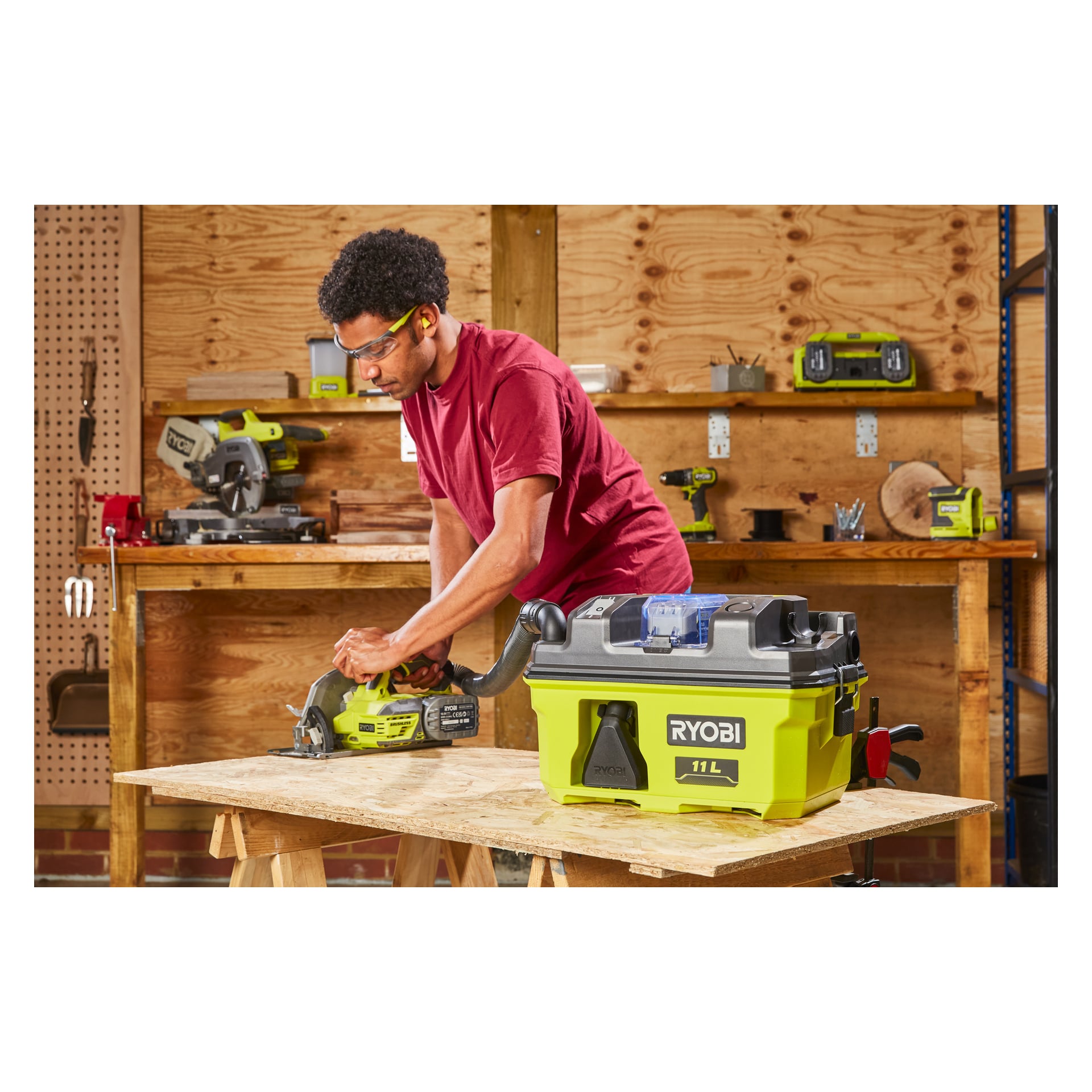 Med vernebriller og hørselvern, betjener man en Ryobi-sirkelsag for å skjære gjennom en treplate i et verksted. På arbeidsbenken og hyllene bak dem kan man se ulike verktøy samt en Ryobi One+ batteridrevet grovstøvsuger RV1811-0.