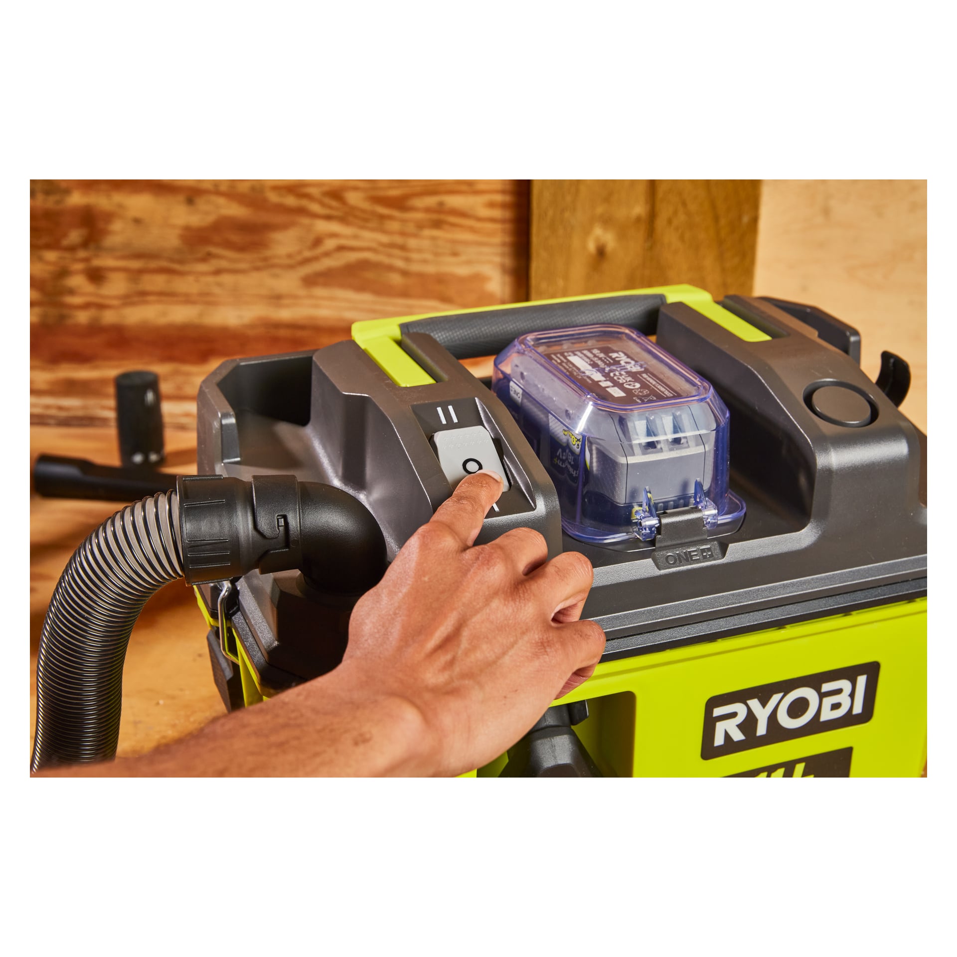 En hånd trykker på av/på-knappen på en Ryobi One+ batteridrevet grov støvsuger RV1811-0, og viser slangen og det gjennomsiktige batterirommet. Vakuumet hviler på en treflate.