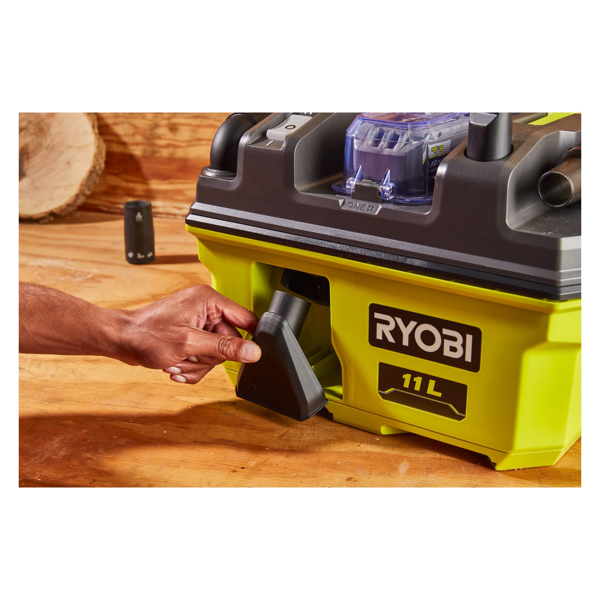 En persons hånd fester en slange eller dyse til en delvis synlig grønn og svart Ryobi One+ batteridrevet grovstøvsuger (RV1811-0) plassert på en treoverflate. Fokuset er på forbindelsesområdet og hånden i aksjon.