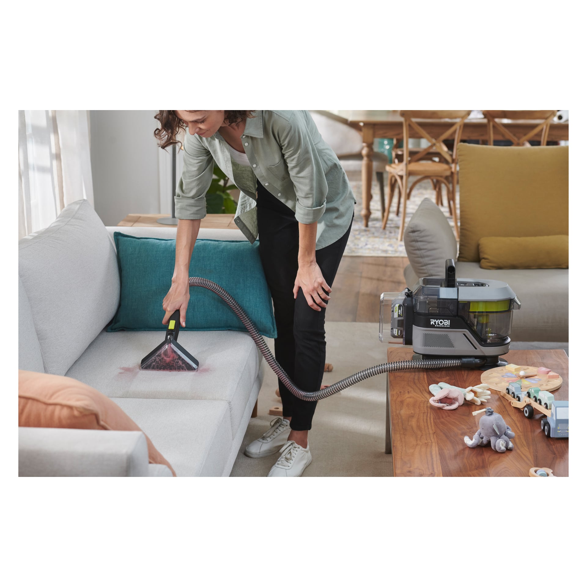 Noen renser en flekk på en lys grå sofa i stua med en Ryobi One+ Swift Clean RDC18BL batteridrevet flekkfjerner. I forgrunnen står det et salongbord dekket av leker og andre ting.
