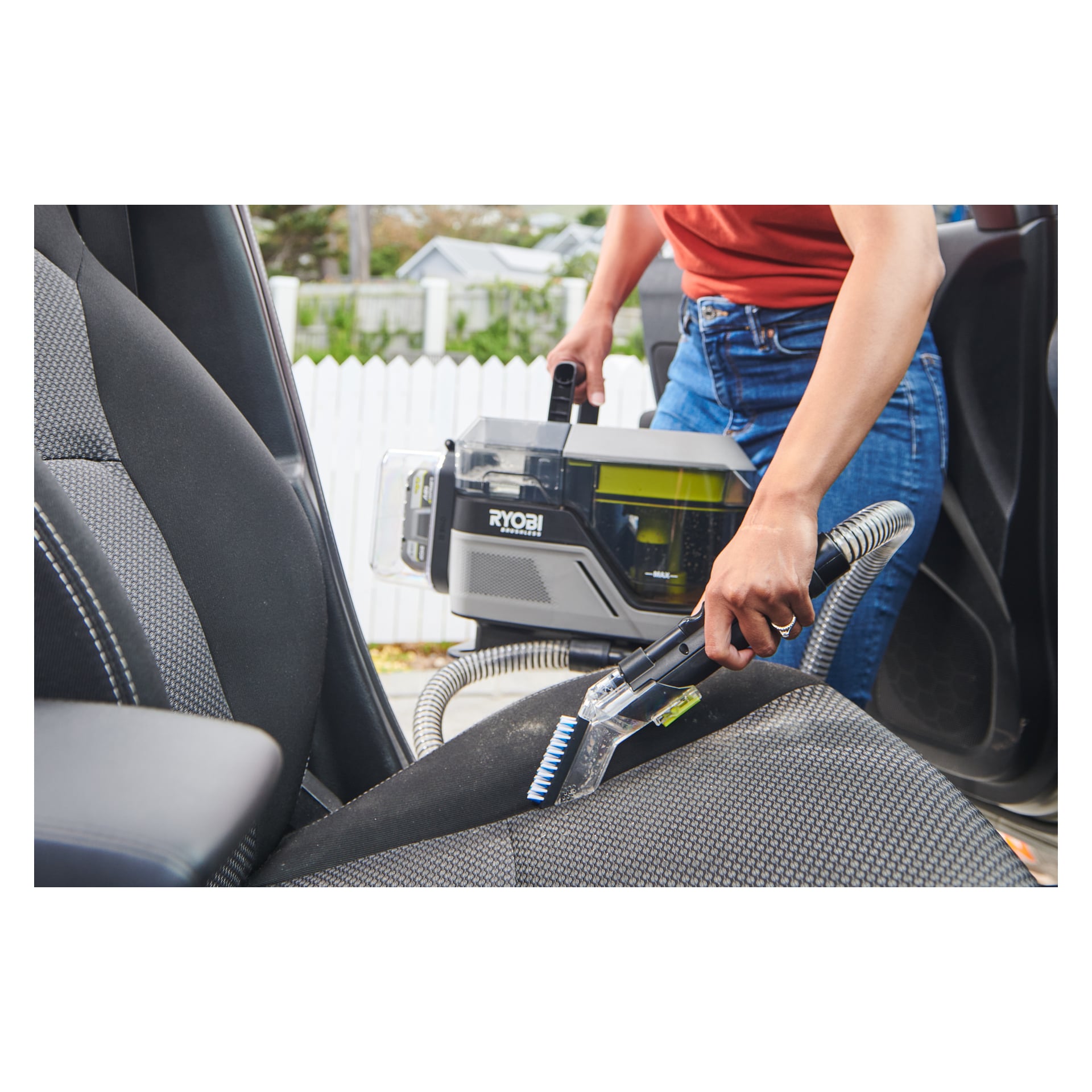 Noen rengjør et stoffsete i en bil med en Ryobi One+ batteridrevet flekkfjerner Swift Clean RDC18BL utstyrt med slange og børste. Personen jobber gjennom den åpne bildøren, og det er et hvitt gjerde i bakgrunnen.