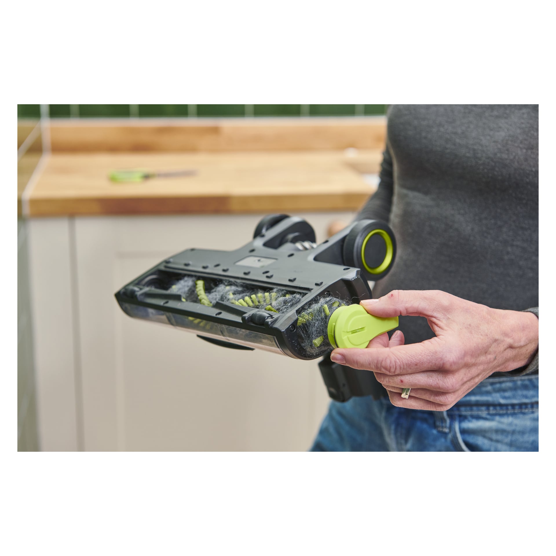 Joku poistaa Ryobi One+ Upright Vacuum Vacuum Cleaner (RSVS18BL-0) -harjanpään päätyä poistaakseen pölyn ja hiukset harjasta. Taustalla näkyy keittiötaso ja seinä, jossa on vihreitä laattoja.
