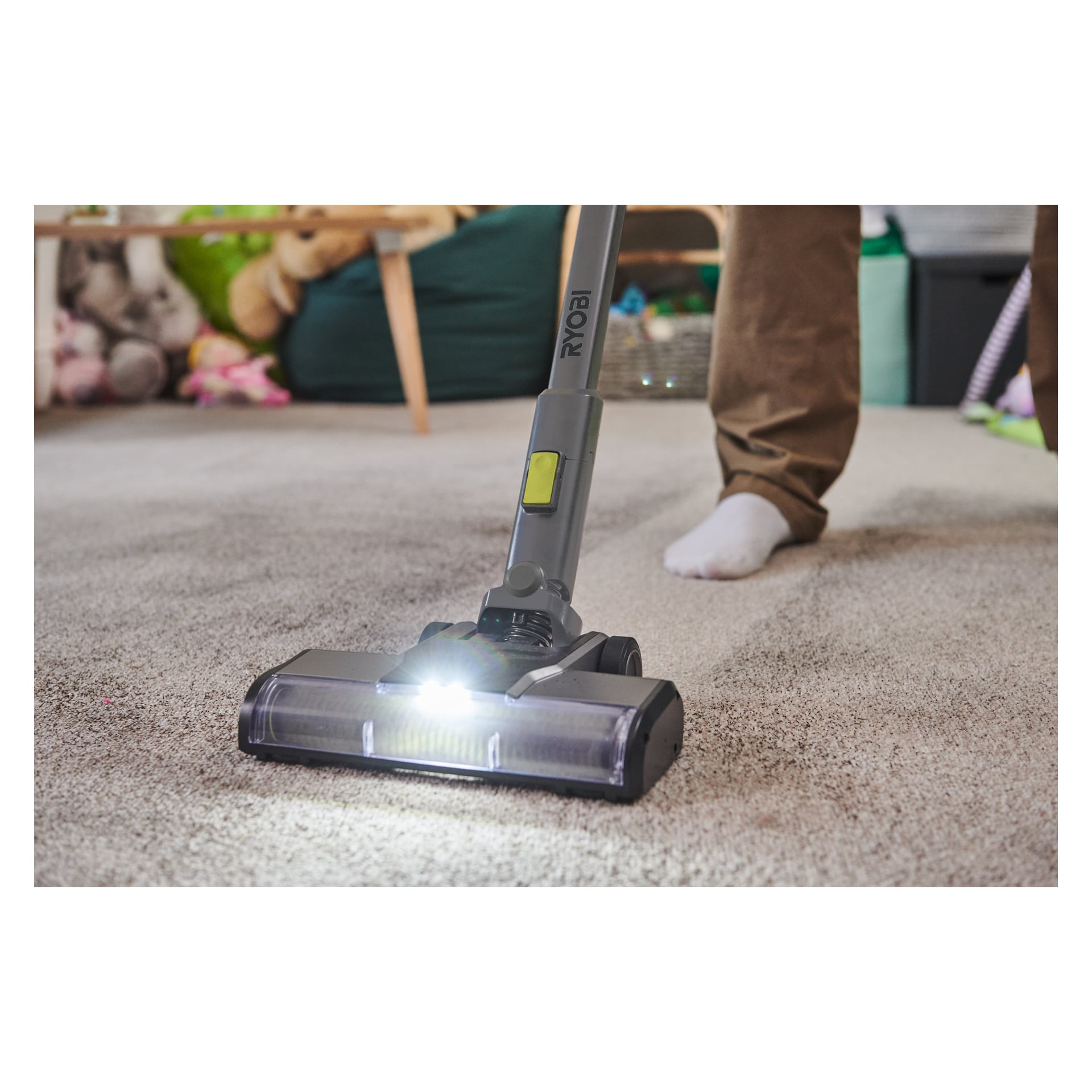 Joku imuroi mattoa Ryobi One+ Upright -pölynimurilla (RSVS18BL-0), jossa on harjaton moottori ja valaistu ajovalo. Taustalla näkyy leluja, kori ja vihreitä huonekaluja.