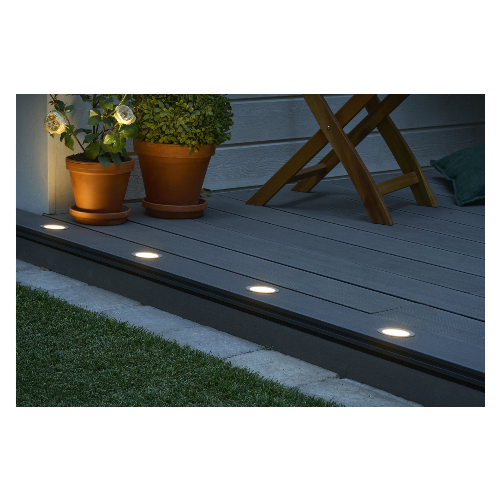 En grå träplatta upplyst av Anslut Decklights Connectable System 4-pack, med runda inbyggda LED-lampor längs kanten. Två orangea krukor med gröna växter, en trästol och gräs finns nära en vit husvägg i bakgrunden.
