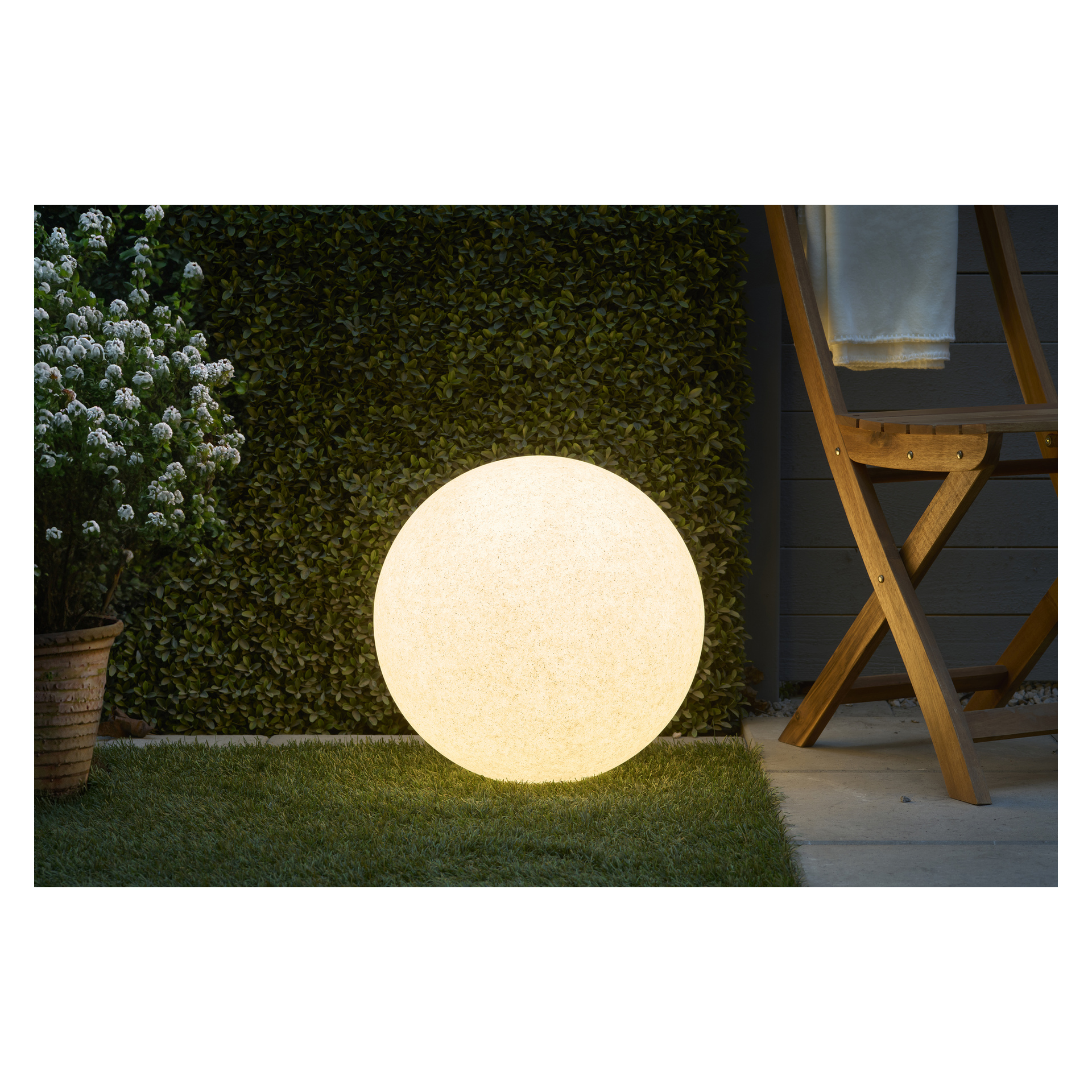 En lysande Anslut LED-svävande sfär från Connectable System vilar på gräs bredvid en trästol och en krukväxt med vita blommor, placerad mot en grön häck.