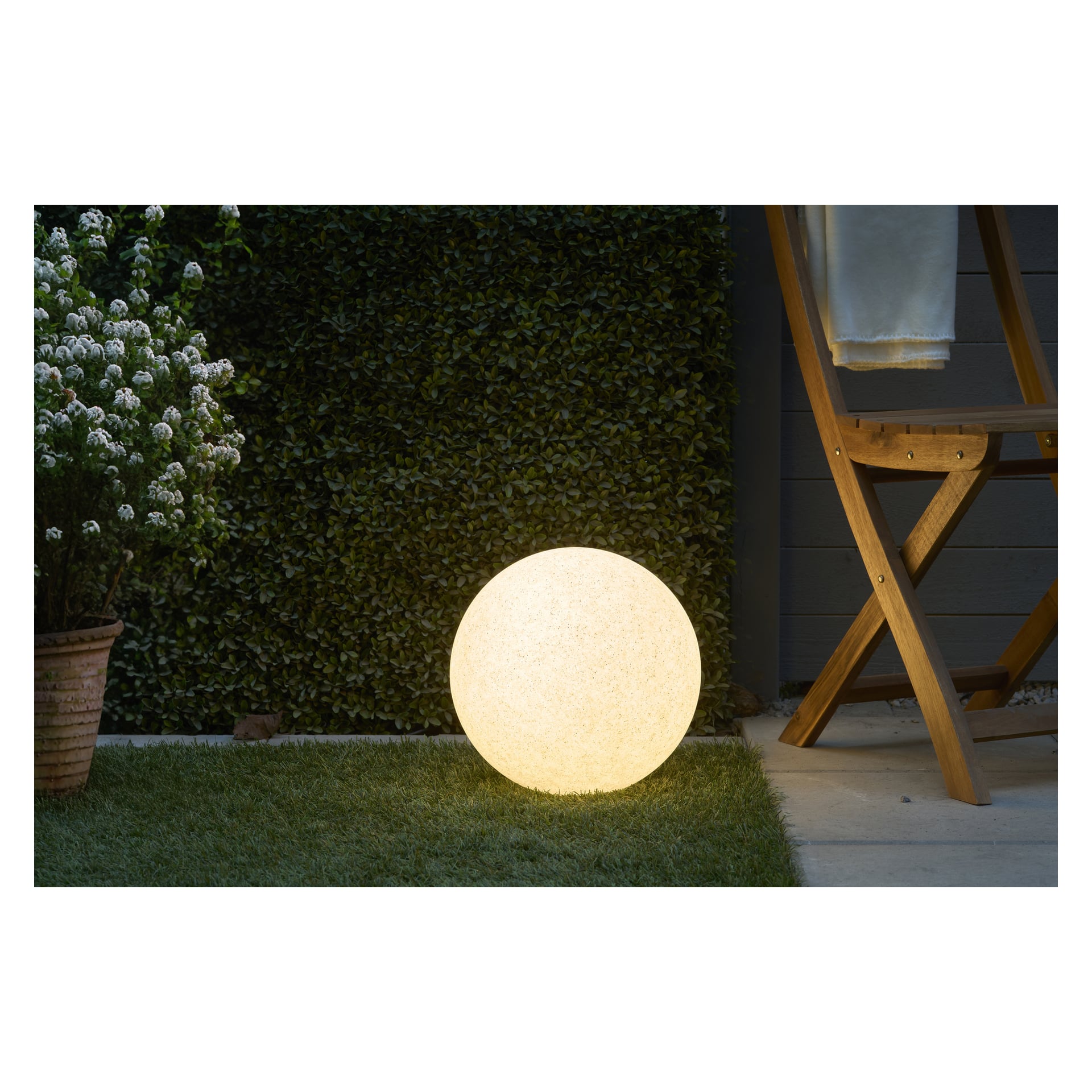 En lysande Anslut LED-svävande sfär från Connectable System-kollektionen står på gräset bredvid en krukväxt, en häck och en trästol täckt med en vit duk.
