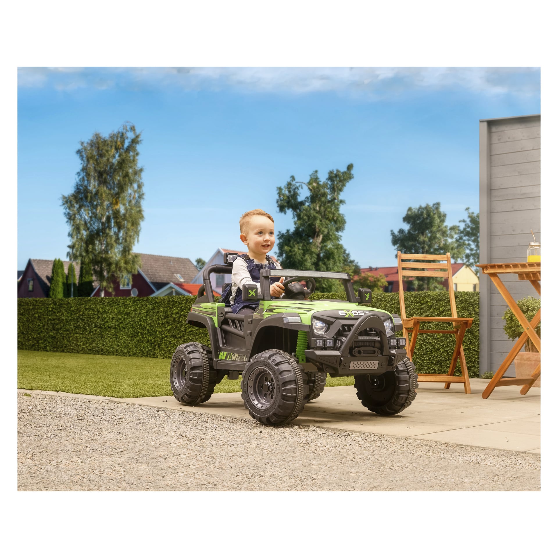 Pieni lapsi istuu Exost ERO Jeep -akkukäyttöisessä autossa patiolla lähellä nurmikkoa. Lähellä on puisia tuoleja ja pöytä, taustalla taloja, puita ja kirkas sininen taivas.
