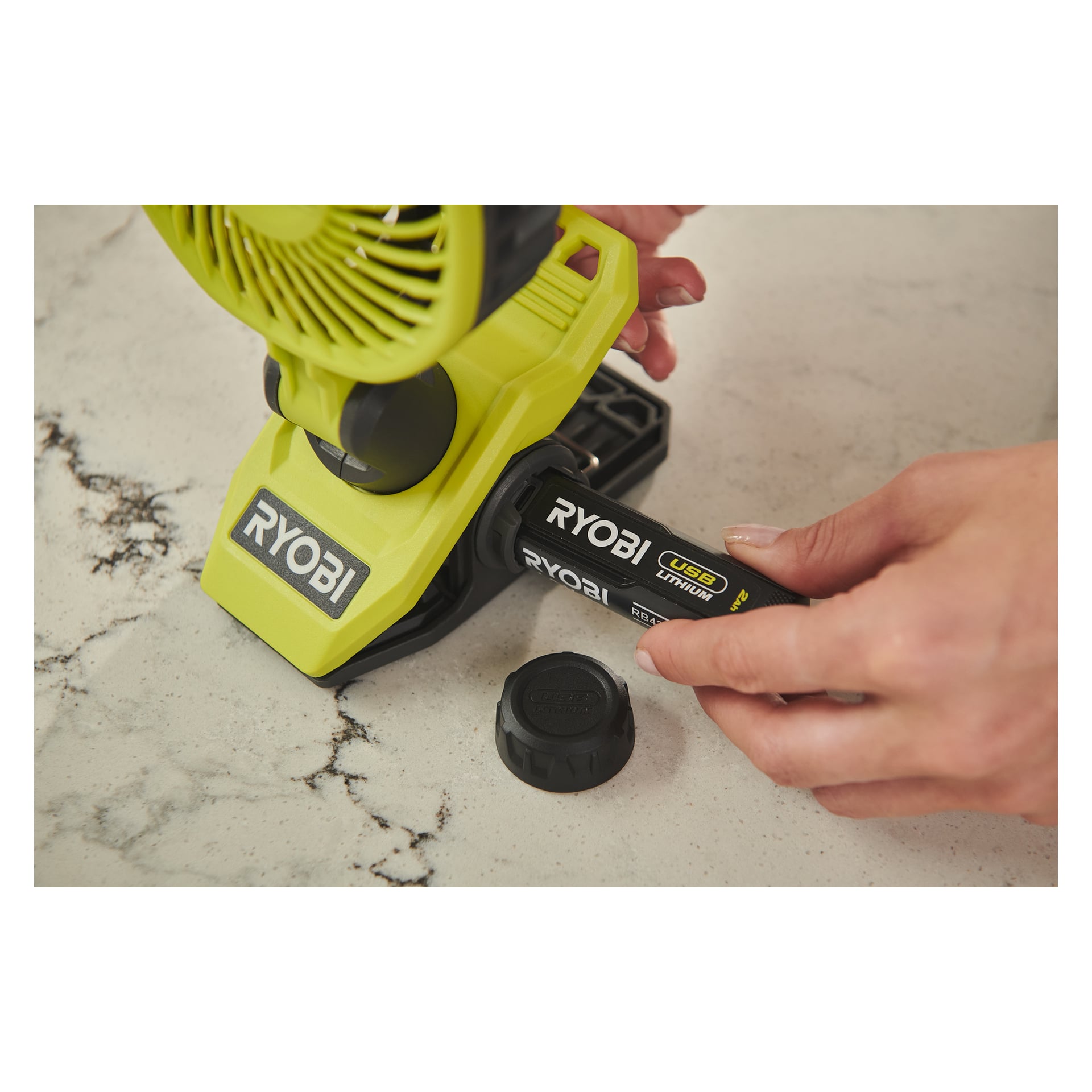 Ktoś wkłada baterię litową Ryobi USB do zielonego wentylatora litowego Ryobi USB z zaciskiem (RCF4-0) na marmurowej powierzchni, z czarnym nasadką baterii umieszczoną obok.
