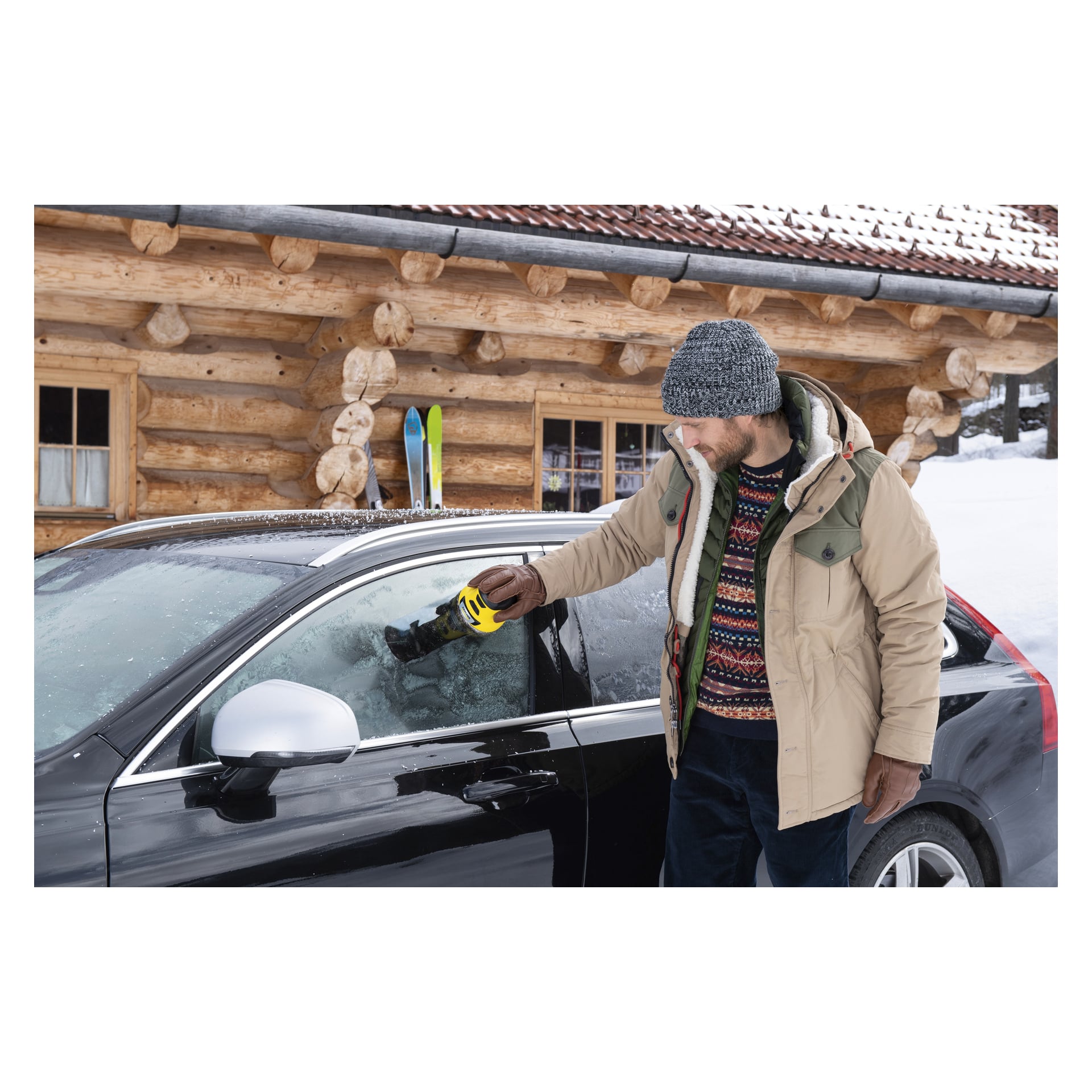 En mann fjerner is fra førersidens vindu på en svart bil ved hjelp av Kärcher Electric isskraper EDI 4, parkert foran en tømmerhytte med snø og ski synlig i bakgrunnen.