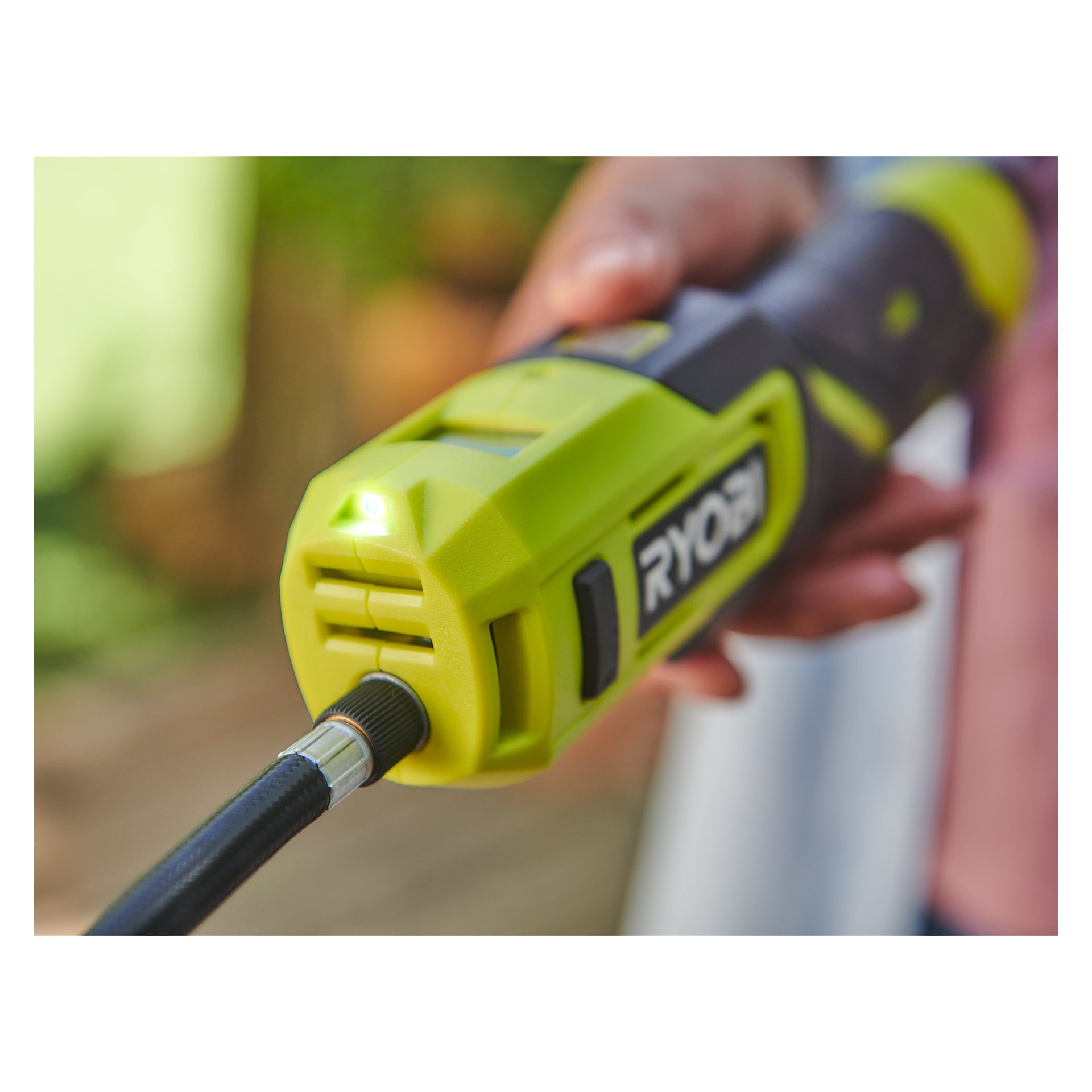Man betjener en grønn og svart Ryobi USB Lithium Battery mini-kompressor RI4-120G med en fleksibel slange utendørs, logoen synlig.