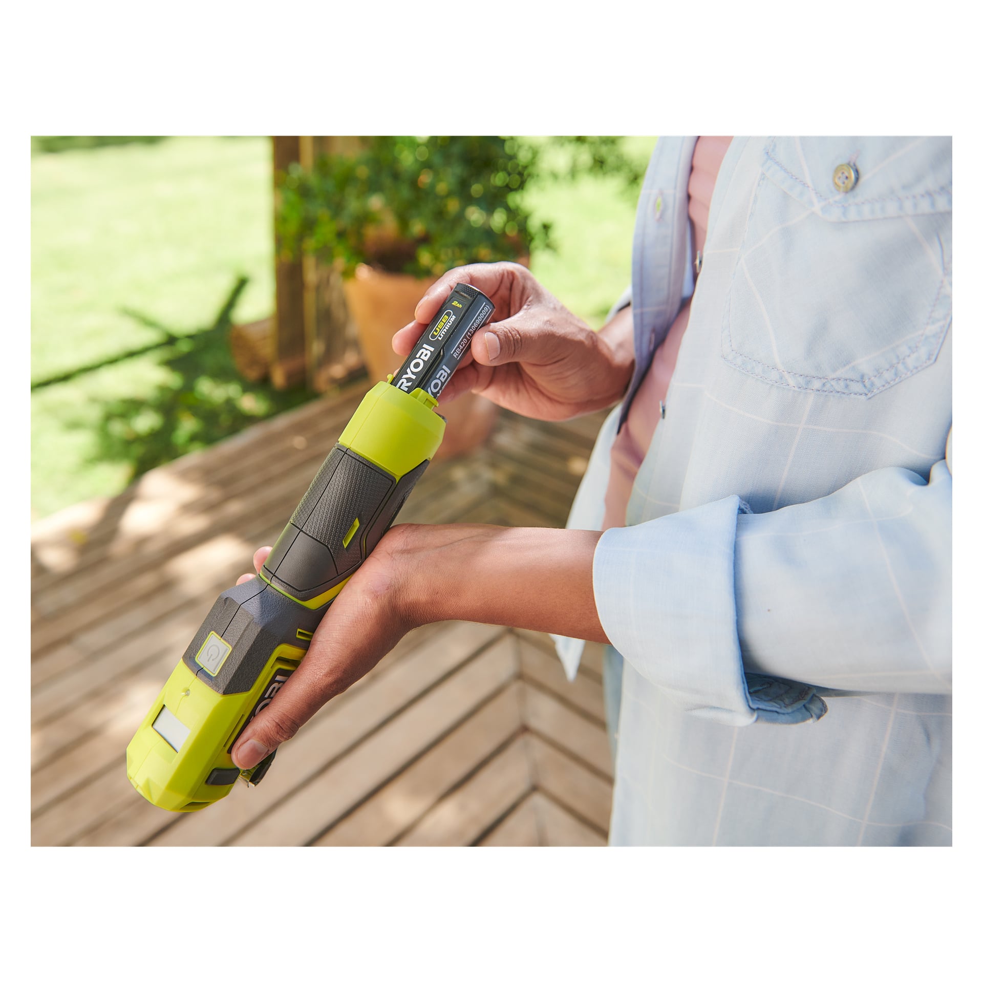 På en treterrasse med frodig grønt og potteplanter, setter en person inn et Ryobi USB Lithium-batteri i RI4-120G mini-kompressoren.