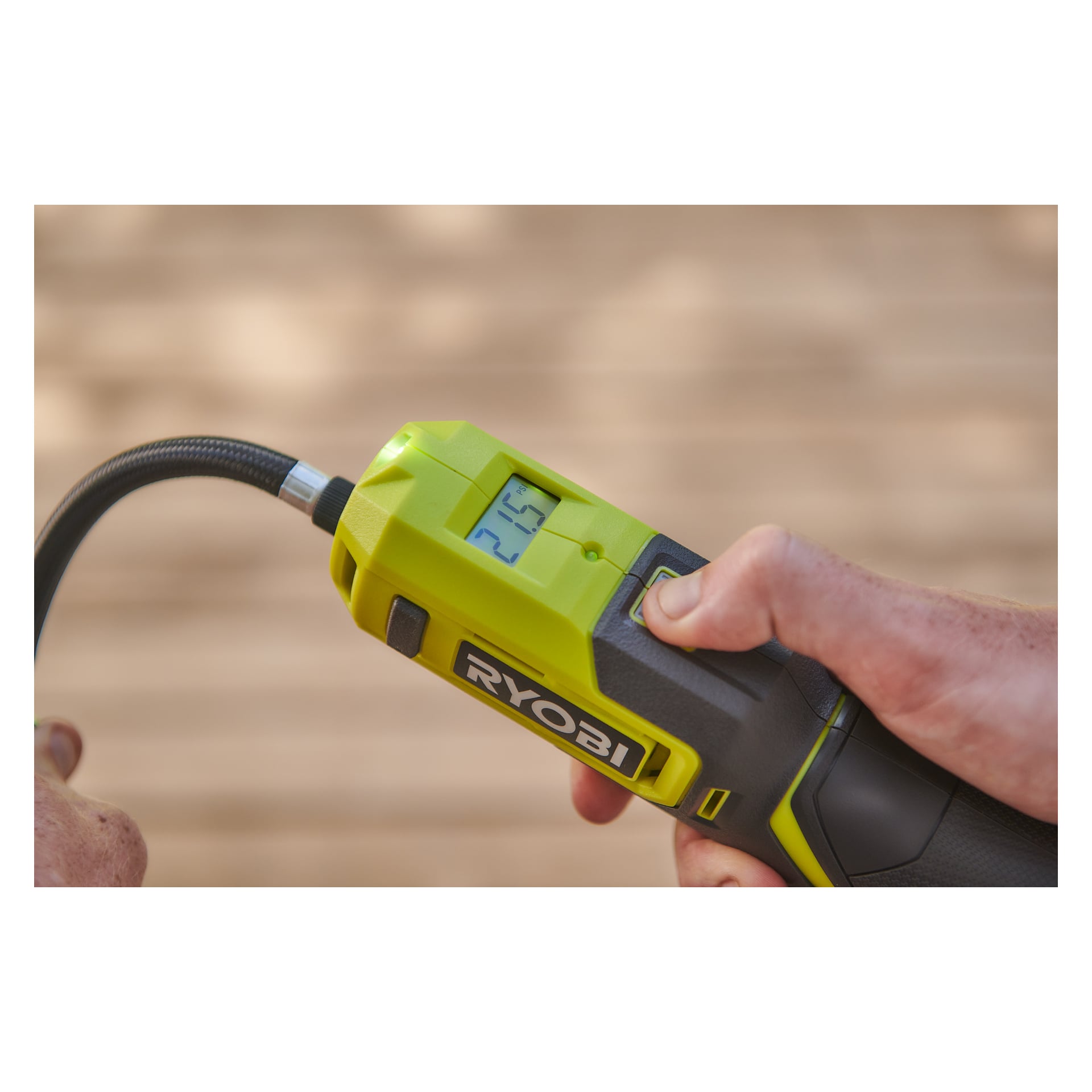 En person holder en Ryobi USB Lithium mini-kompressor RI4-120G som viser en digital skjerm på 21,5, slangen er koblet øverst.