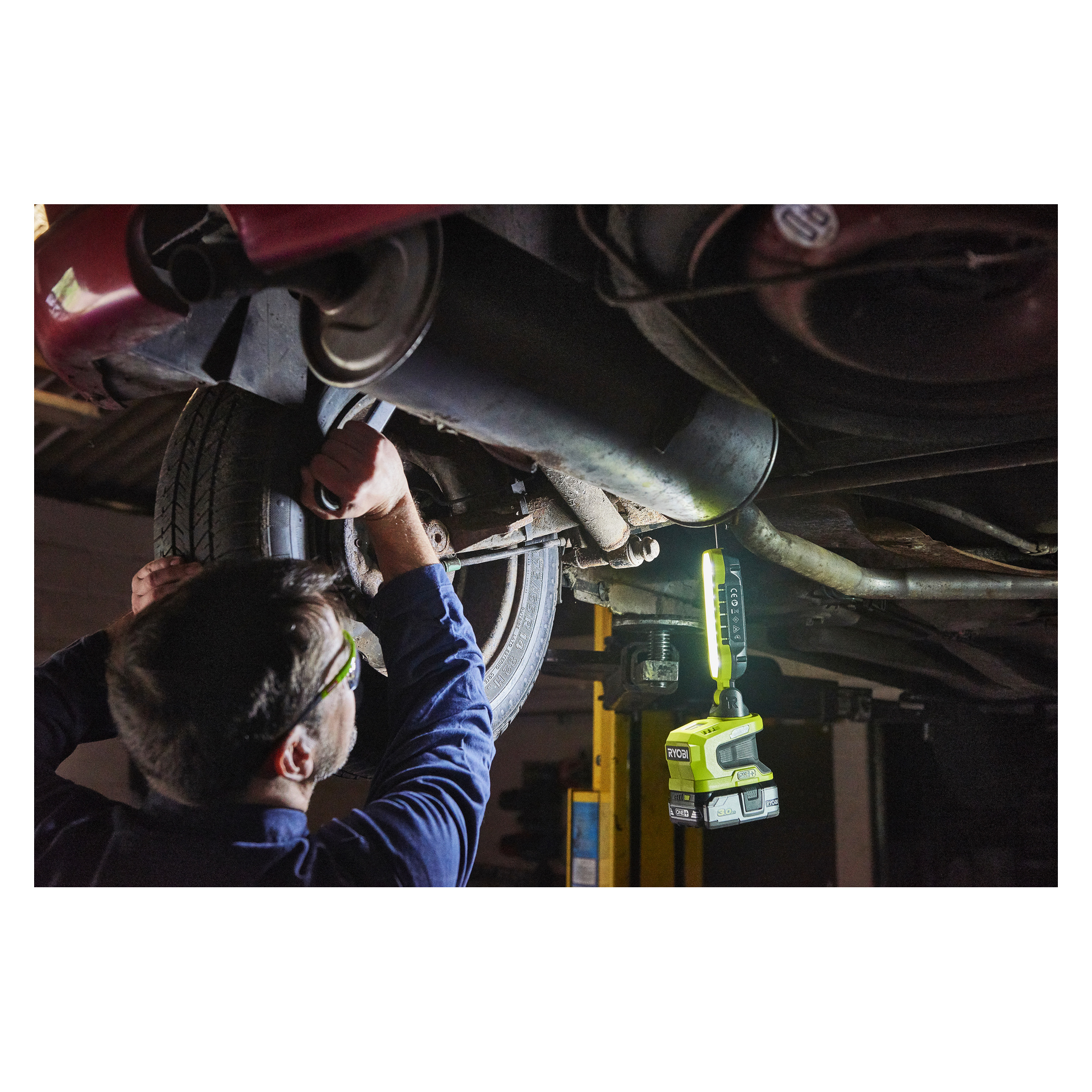 En person med skyddsglasögon använder ett verktyg nära bilens bakhjul, med en Ryobi One+ LED-arbetslampa R18ALP-0 som lyser upp området.