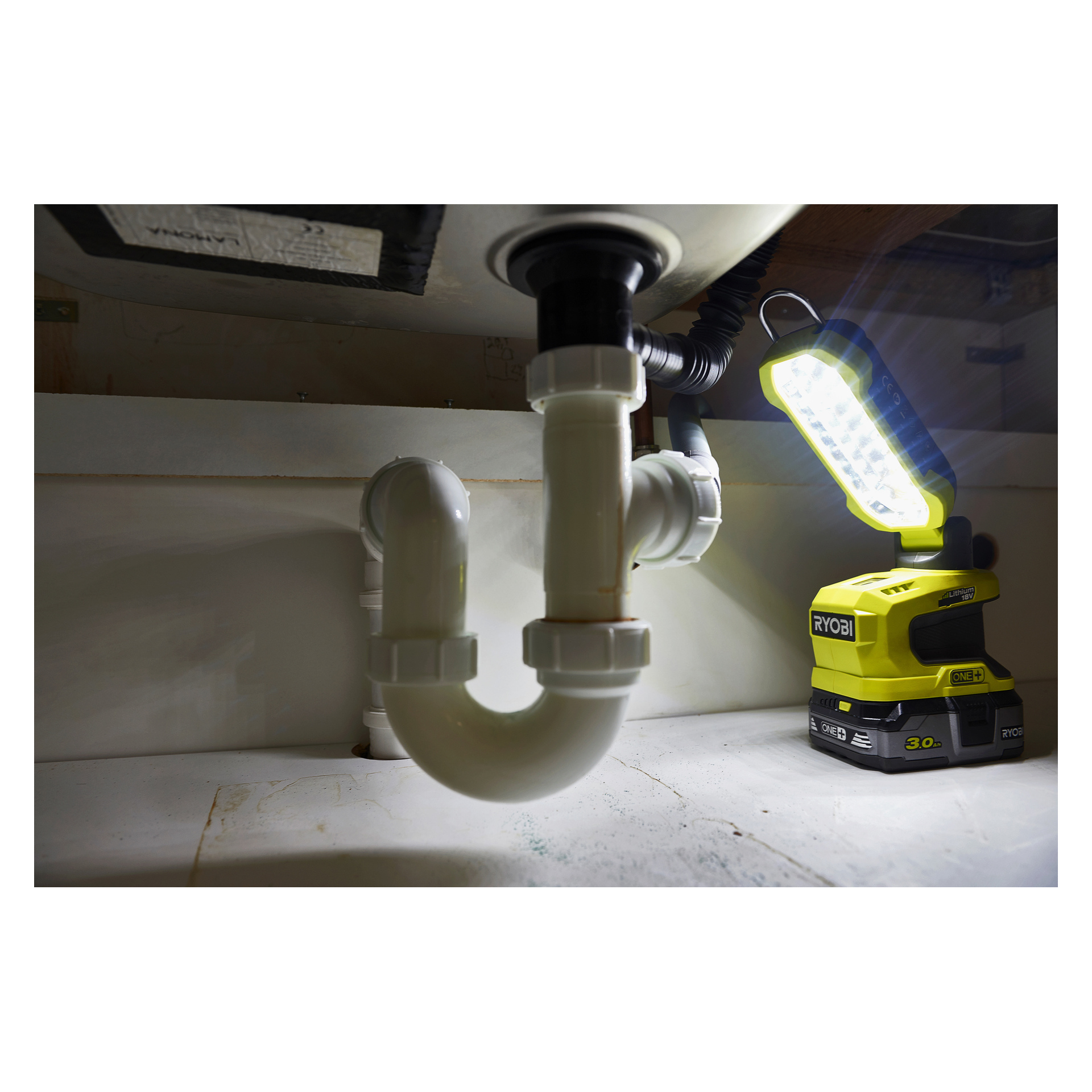 En Ryobi One+ LED-arbetslampa R18ALP-0 lyser starkt upp prydliga vita PVC-rör under en diskho i ett prydligt och organiserat utrymme.