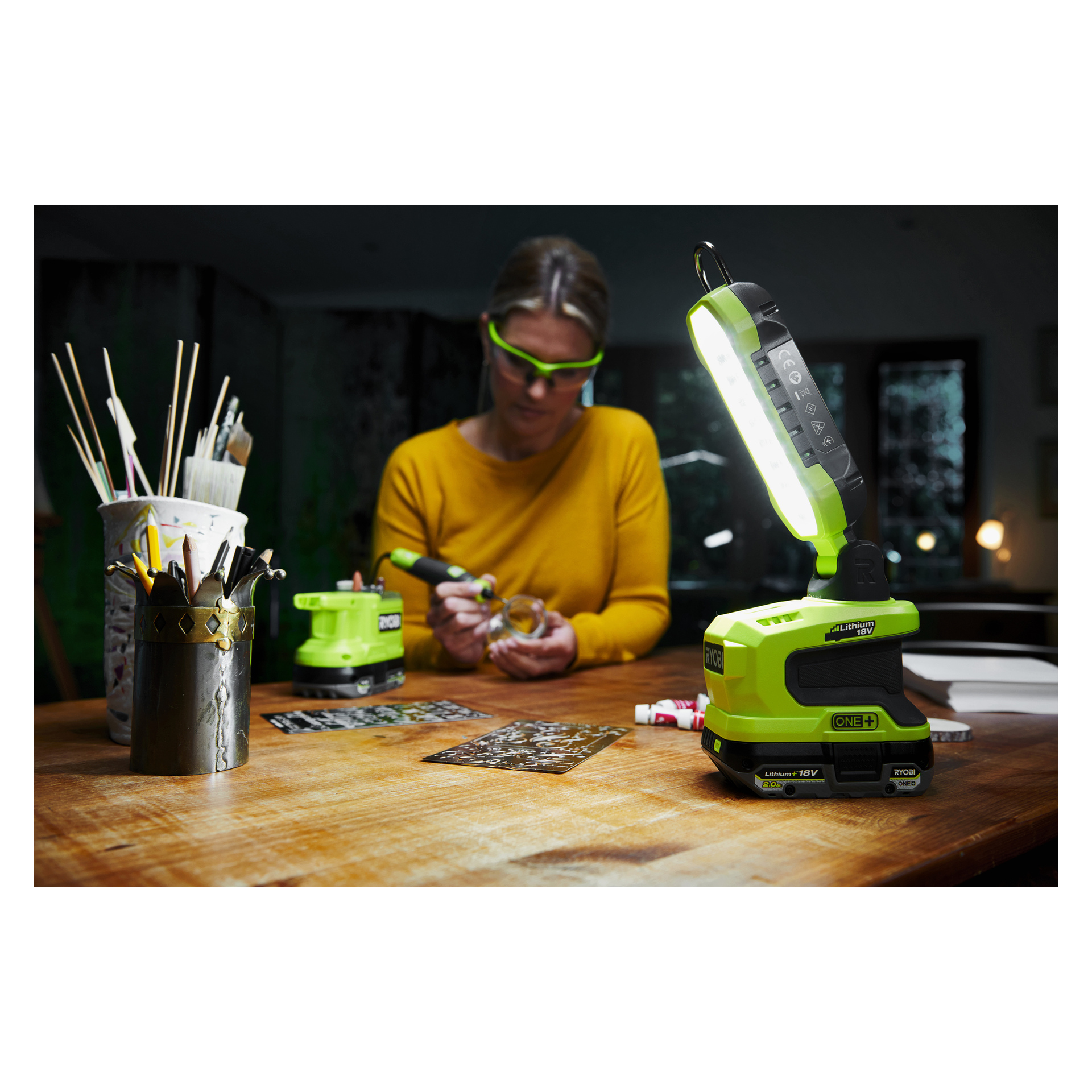 Med skyddsglasögon och gul skjorta använder man en Ryobi One+ LED-lampa vid ett pysselbord med penslar och färger.