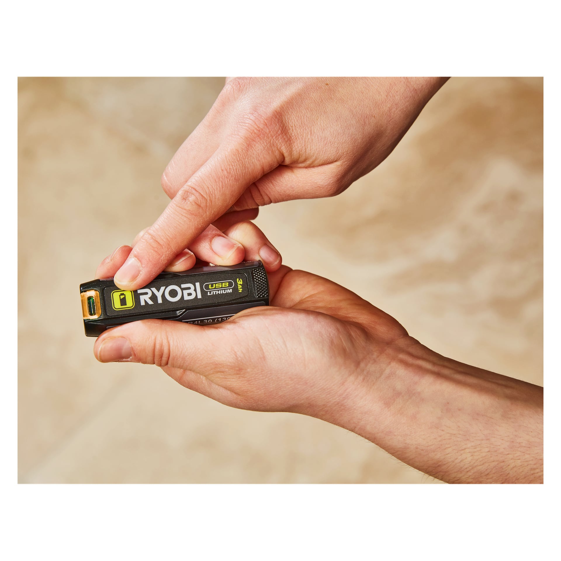 Noen holder en Ryobi USB Lithium Battery RB4L30 i den ene hånden, trykker på knappen med den andre, bakgrunnen er mykt ute av fokus.
