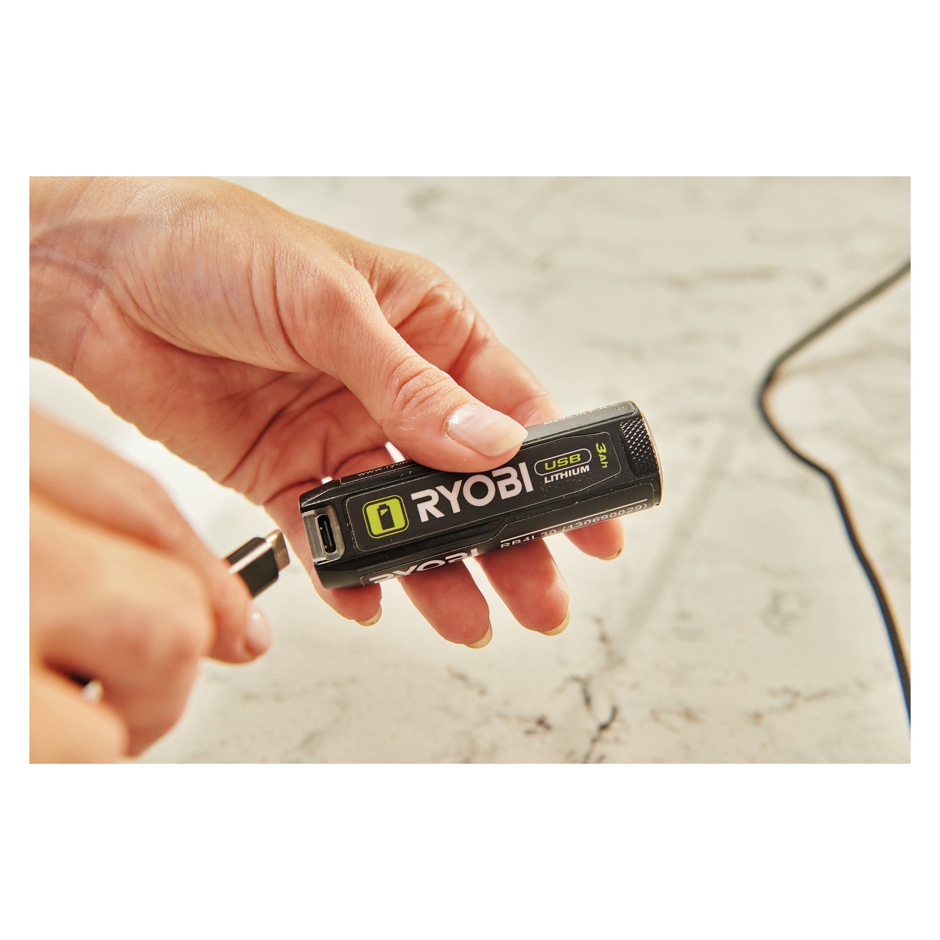 En hånd griper et Ryobi USB Lithium Battery RB4L30 mens en annen hånd justerer en USB-kabel for å koble til, plassert mot en flekkete overflate.