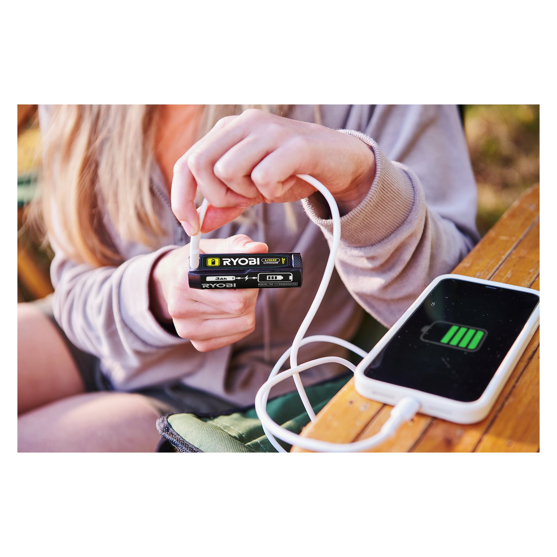 Sittende ved et utebord i tre bruker en person en hvit kabel til å lade telefonen fra et Ryobi USB Lithium Battery RB4L30.