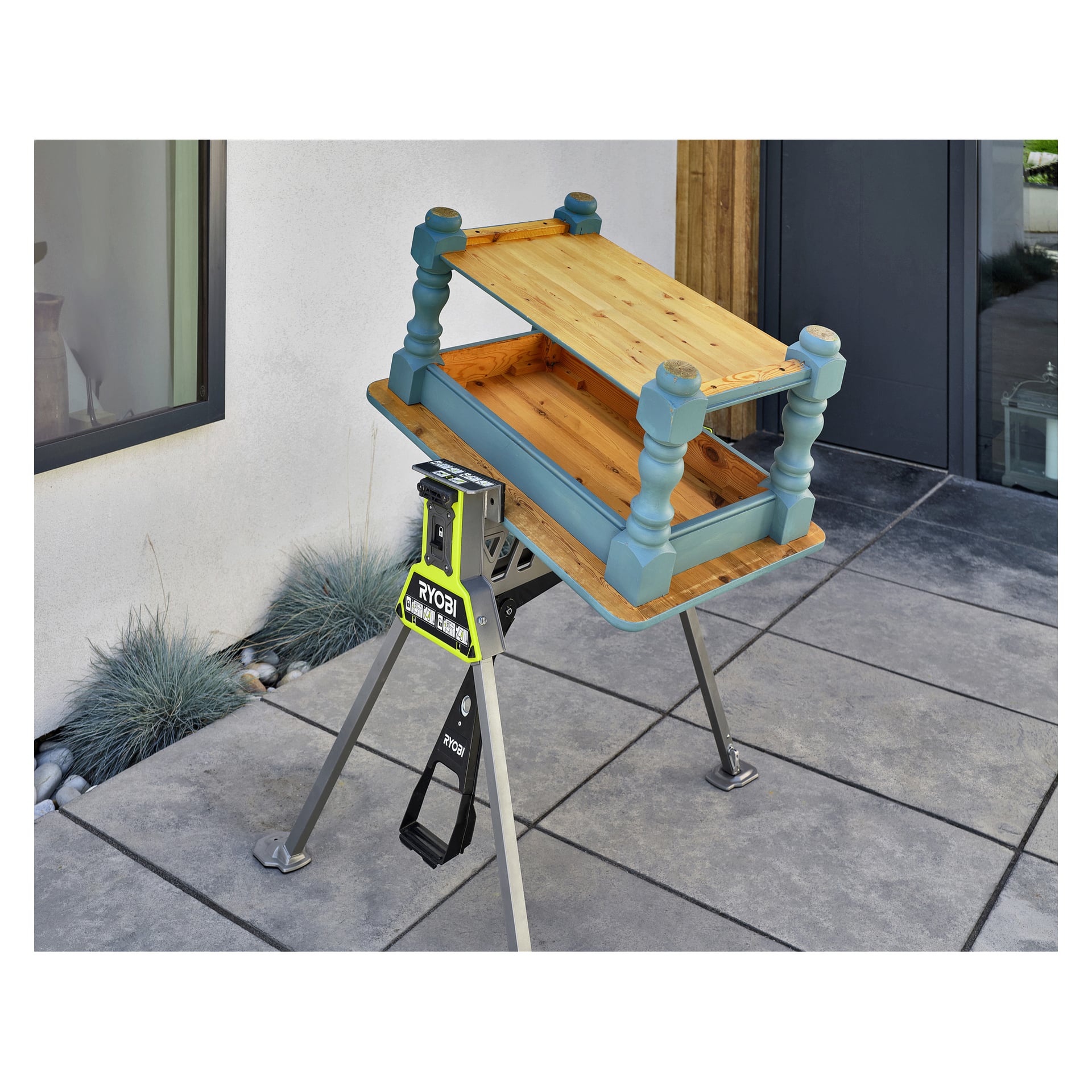 Ett litet blått och träbord står upp och ner på en Ryobi One+ hopfällbar arbetsbänk med klämma, utanför ett modernt hus nära växter.