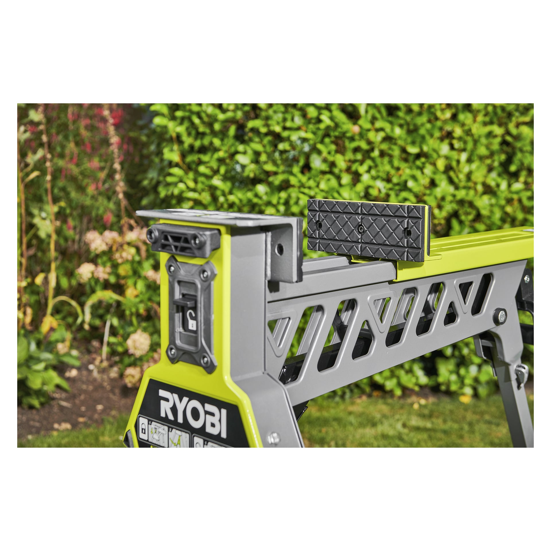 Närbild av en Ryobi One+ hopfällbar arbetsbänk RSTSJ150 med klämma, grå och gul, på gräs, grön växtlighet i bakgrunden.